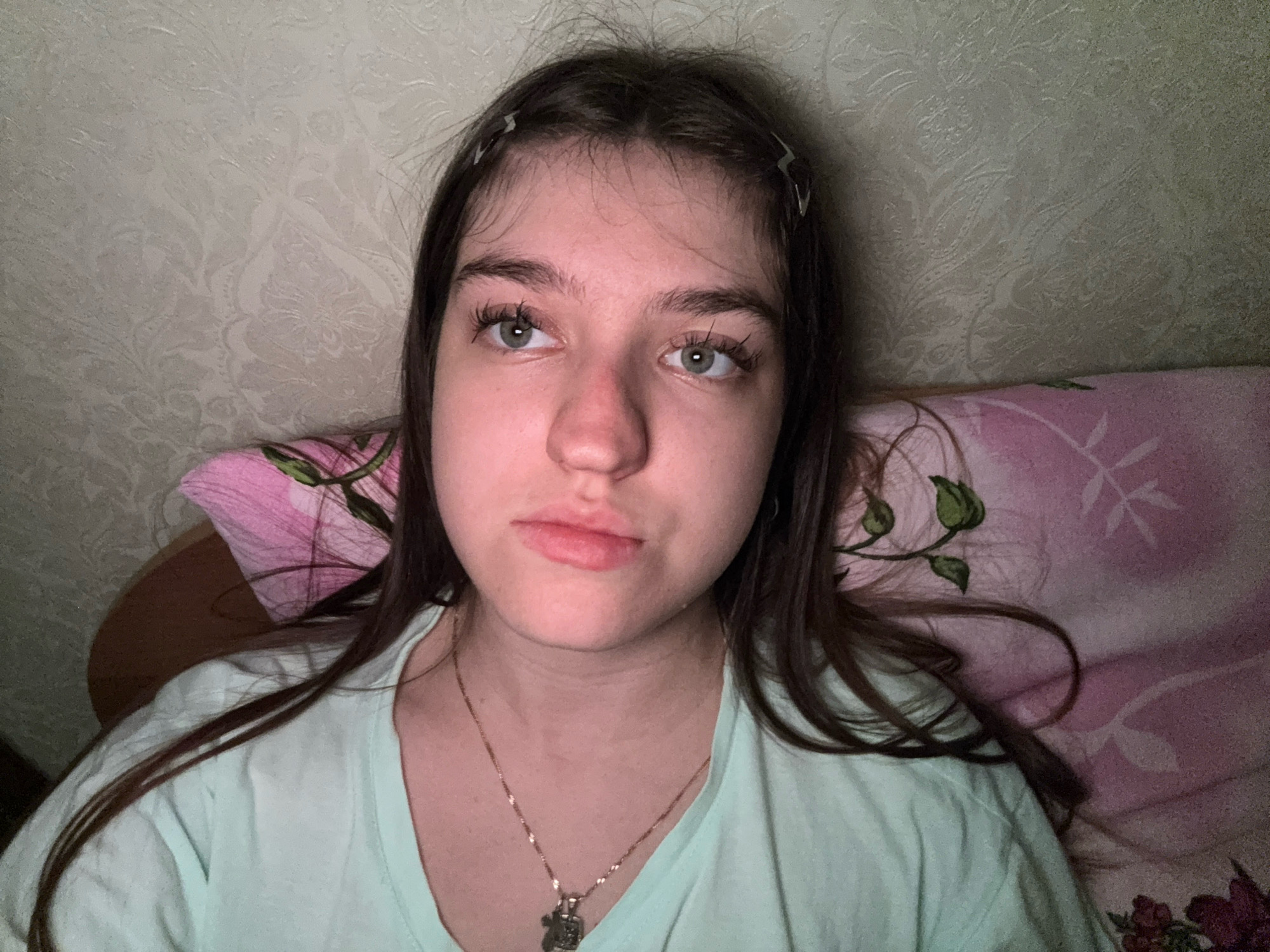 A medium shot shows a young woman looking upwards and to her left, with her head slightly tilted. She has long, dark brown hair, some of which is pulled back with hair clips. Her eyes are green, and she is wearing a light green t-shirt with a gold necklace featuring a pendant. Her lips are slightly parted, and her mouth is turned in a slight smirk or contemplative expression. The background appears to be a textured wallpaper, and behind her on the right is a pink pillow with a floral pattern. The lighting suggests an indoor setting, possibly a bedroom, with the light source coming from above and slightly to the left, casting a shadow on the right side of her face. There are no visible indicators of the city of Zavolzhye, Russia.