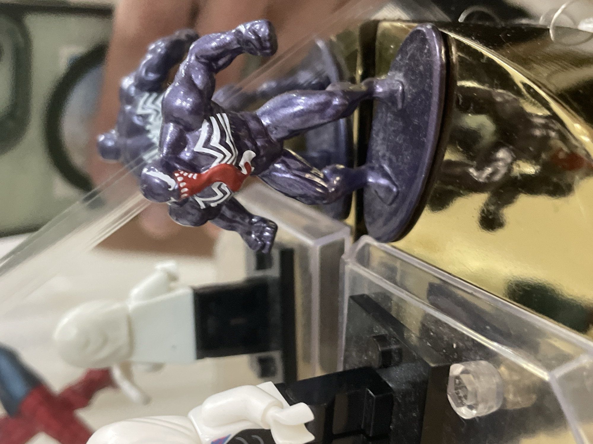The image, taken in Ihtiman, Bulgaria, captures a close-up of several action figures arranged within a clear plastic and reflective golden display case. The most prominent subject is a dark purple (or black) Venom miniature, dynamically posed mid-action with clenched fists and bent limbs, as if lunging forward. The figure features its iconic white spider emblem on the chest and back, and a hint of red is visible near its head, likely representing its tongue. It stands on a circular dark purple base, which is integrated into the display structure.
A highly reflective golden surface behind the Venom figure creates a strong, distorted reflection of the figure, adding a sense of depth. Below the main figure, partially visible through clear acrylic panels, are other miniature figures. One is a white, simplified block-style figure, resembling a Lego character, standing on a black base. Further to the bottom left, a red and blue figure, possibly Spider-Man, is indistinctly visible. The display case consists of clear plastic panels, black structural elements, and a visible clear hexagonal peg connector. The background is out of focus and nondescript, suggesting an indoor setting. No discernible text is present.