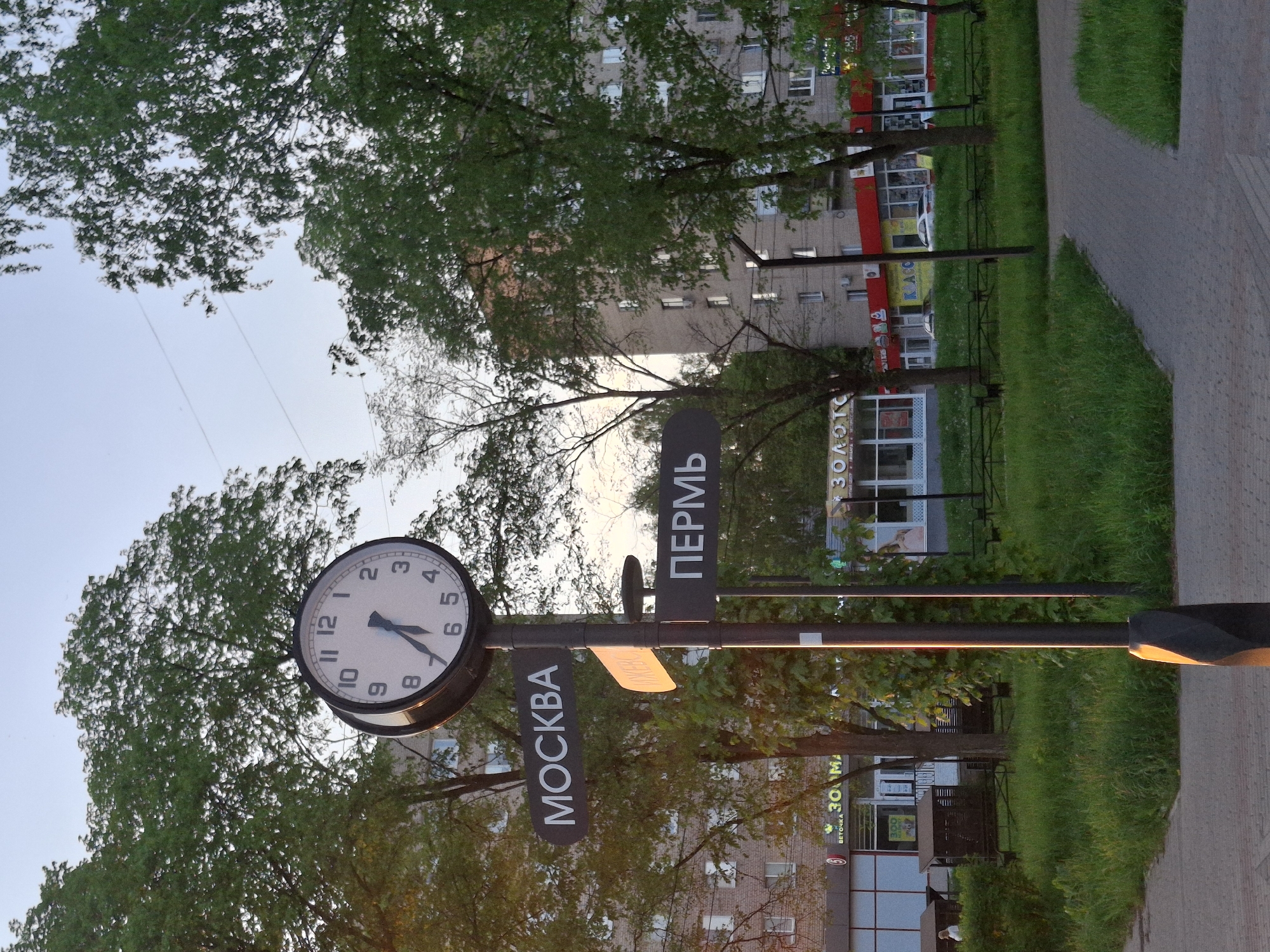 The image depicts a street scene in the city of Chaikovsky, Russia, featuring a clock post with signs indicating directions to Moscow and Perm. The clock face shows the time as approximately 5:35. The signage is in Russian Cyrillic script. The clock post is surrounded by trees and greenery, with a sidewalk and grass visible in the foreground. Buildings are visible in the background, including structures with commercial establishments on the ground floor. The scene appears to be captured during daylight hours, with the lighting suggesting late afternoon or early evening.