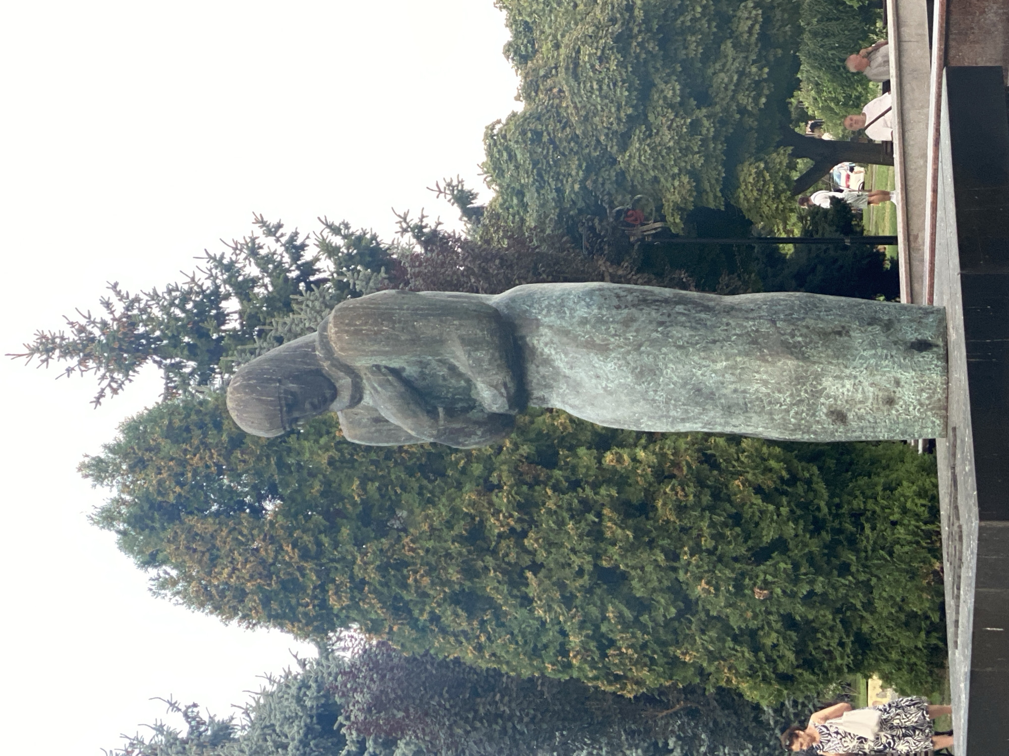 The image depicts a bronze statue of a woman, possibly a mother holding a child, positioned in an outdoor setting with lush greenery. In the foreground, the base of the statue is visible, a dark, rectangular structure. The statue itself is tall, with a weathered patina, and the woman's head is bowed. Behind the statue, various trees create a dense backdrop.
To the left of the statue, a woman in a black and white patterned dress walks with a white bag. Further back and to the right, several people are seated on a bench, observing the surroundings. One man wears a white t-shirt, another a light-colored shirt. Another individual, a man in a white shirt and shorts, stands in the background.
The overall environment suggests a public park or plaza, likely in Chișinău, Moldova. The statue is a prominent feature, surrounded by mature trees and accessible to the public, indicated by the presence of people. The sky is overcast, contributing to a soft, diffused light across the scene.