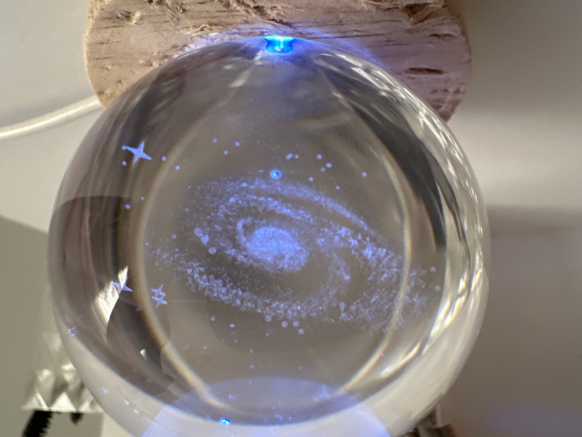 A clear glass orb with a galaxy design sits on a cork coaster. The orb is illuminated from within by blue light, highlighting the swirling galaxy pattern and small blue dots resembling stars. A white cable is visible to the right of the orb. In the background, out of focus, is a white object with a faceted design and what appears to be dark bristles. The scene is well-lit and appears to be indoors on a white surface.