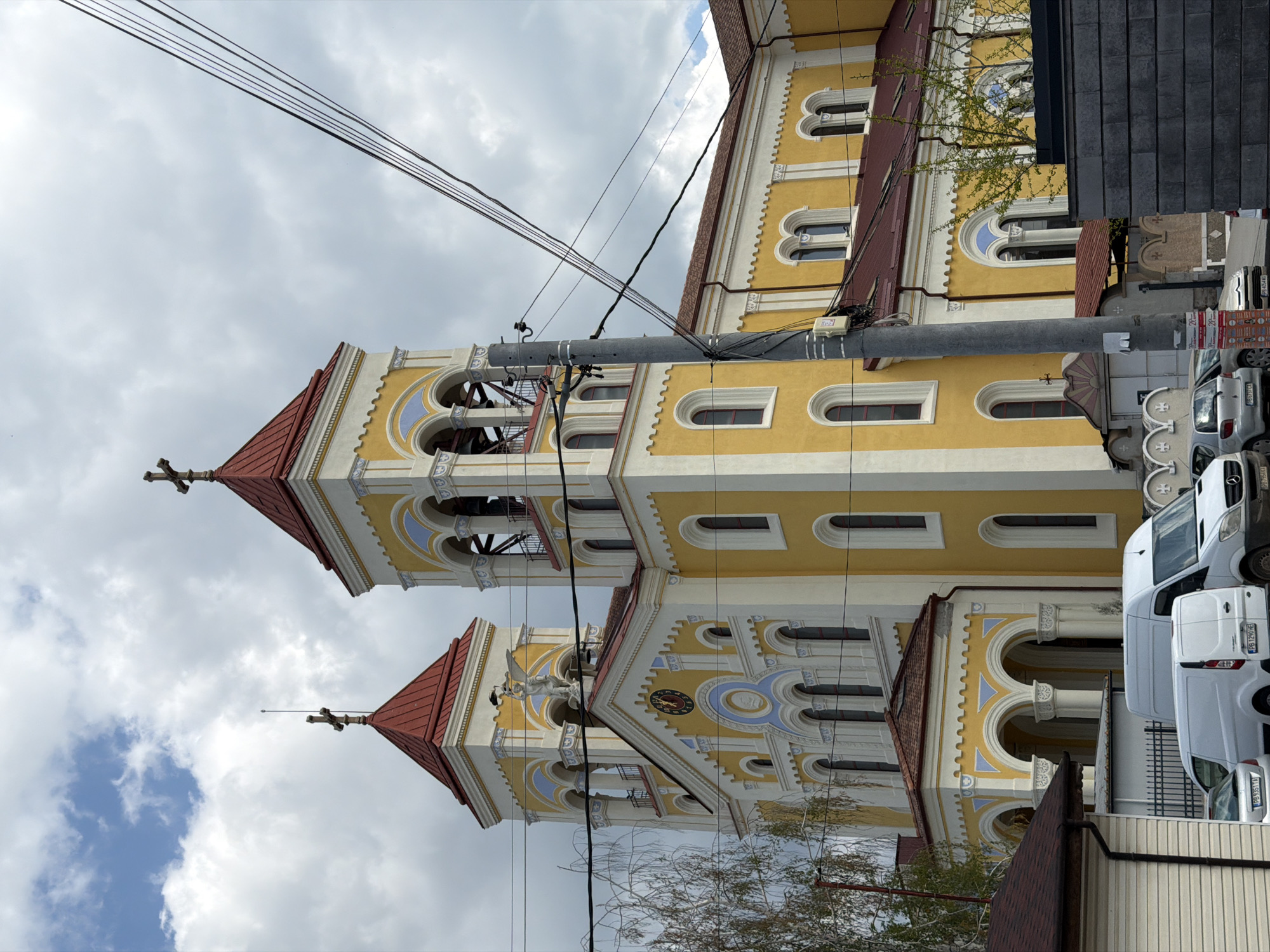 The image, when viewed upright, depicts an ornate church building in Rakovski, Bulgaria, under a cloudy daytime sky. The church features a vibrant yellow facade with extensive white trim and light blue decorative accents, including numerous arched windows and ornamental cornices. Two prominent bell towers, each topped with a steep red conical roof and a cross, rise towards the sky. A statue of an angel or saint is visible near the peak of one section of the church, and a clock face with Roman numerals is integrated into another, though the time is illegible.
The foreground and midground are significantly intersected by numerous black overhead utility wires and a sturdy grey utility pole, which partially obscure the church's architecture. On the right side of the image, several vehicles are parked along the street. These include two white Ford Transit-style commercial vans and a silver car. Visible Bulgarian license plates on these vehicles, such as "PB 2596 AB," "PB 9313 TA," and "PB 4241 AB," indicate a registration from Plovdiv Province, consistent with the location context.
A section of a dark grey, possibly tiled, building or wall is visible on the far right. Scattered tree branches, some bare and some with burgeoning green leaves, are present. The scene is static, with no human activity or specific event taking place, presenting a street view centered on the distinctive church.