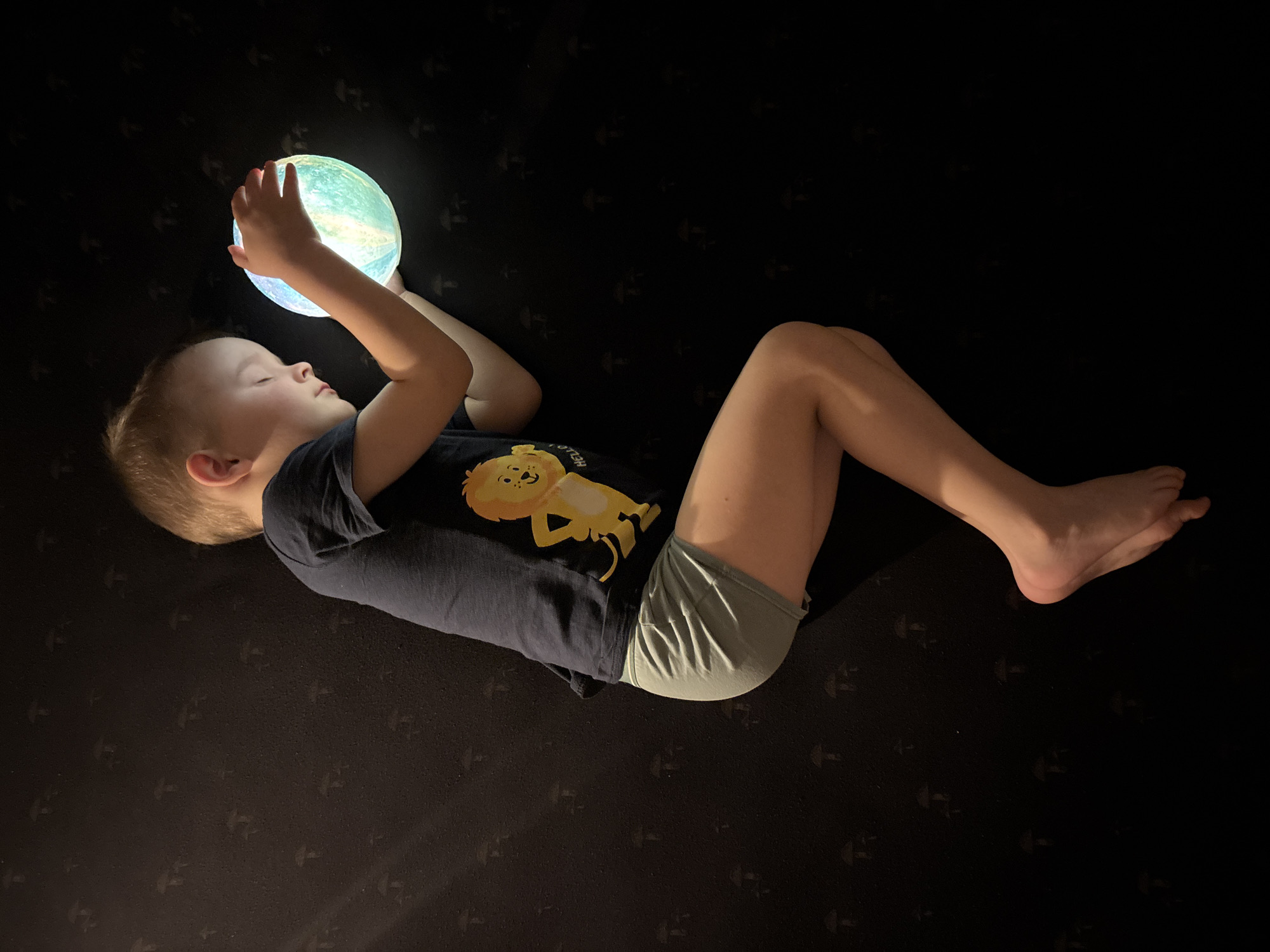 A young boy is captured lying on his back on a dark, subtly patterned surface, holding a glowing, multi-colored spherical object (predominantly blue, green, and white) above his chest. His eyes are closed or nearly closed, and his expression appears peaceful and serene, suggesting he is either asleep, deeply contemplative, or mesmerized by the light.
The boy is dressed in a dark blue t-shirt featuring a cheerful yellow cartoon lion, with the word "HELLO" partially visible upside down near the bottom of the design. He is also wearing light green shorts. His arms are bent, cradling the luminous sphere, while his legs are comfortably bent at the knees.
The scene is set indoors, likely in a bedroom or a similar personal space, characterized by very low ambient light, implying it is nighttime or a darkened room. The glowing sphere is the primary light source, casting a gentle illumination on the boy and his immediate surroundings. The dark surface beneath him has a recurring, indistinct pattern of small, stylized shapes, which adds subtle texture to the background. The overall mood of the image is tranquil and calm. The image was taken in Chaikovsky, Russia.