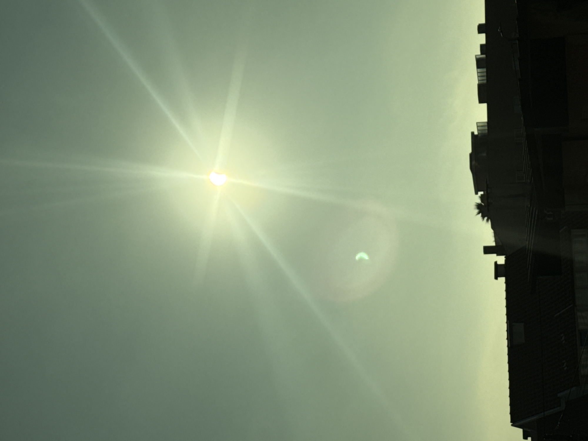 The image shows a partial solar eclipse over the city of Vila Nova de Gaia, Portugal. The sun, partially obscured by the moon, is visible in the upper center of the frame, emitting bright rays of light. Below the sun, silhouetted against the sky, are the rooftops of several buildings. The overall color tone of the image is a hazy green, likely due to atmospheric conditions or a filter used during the photograph. There are no people or discernible actions visible in the image, focusing solely on the celestial event and the urban landscape.