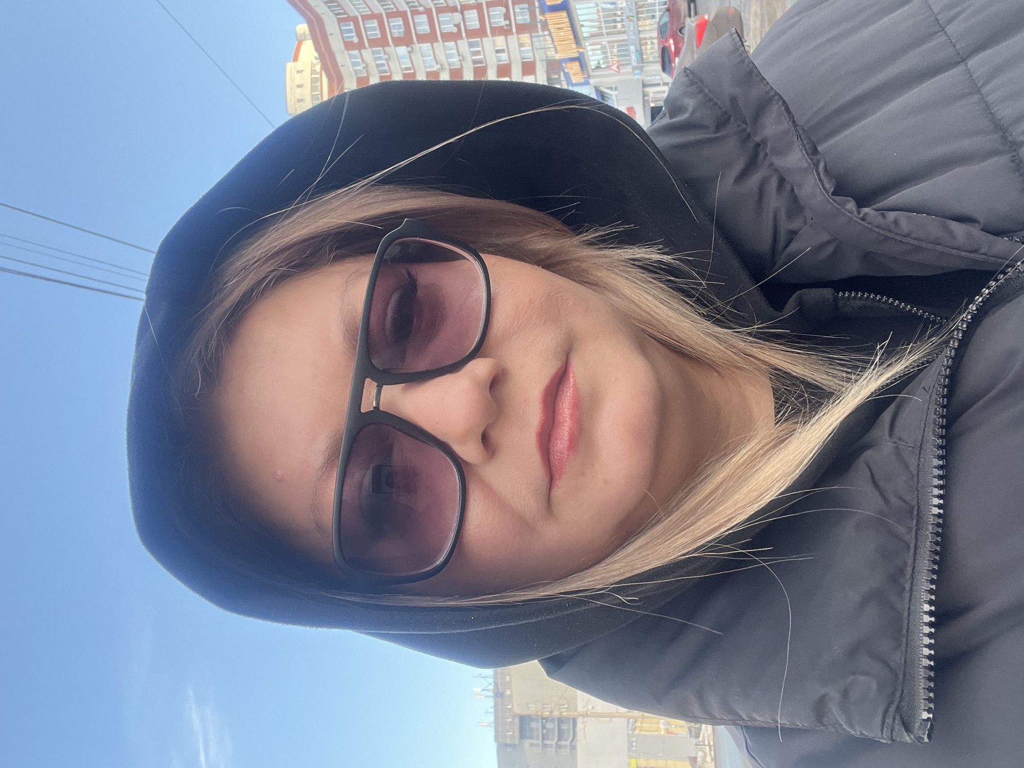 An adult individual, likely female, is captured in a close-up, front-facing self-portrait. She wears dark-framed sunglasses with purple-tinted lenses and a black hooded garment pulled over her head, partially obscuring her light-blonde hair. A black puffer jacket with a visible zipper is worn over a dark inner layer. A small epidermal lesion is present on her forehead, superior to her right eyebrow. Her lips are adorned with pinkish-red lipstick.
The scene is an urban outdoor environment under a clear, bright blue sky. Overhead electrical or communication lines are visible against the sky.
The background identifies the location as Izhevsk, Russia. To the right of the individual, a multi-story residential building with distinct reddish-brown and white facades and numerous rectangular windows is prominently visible. Below this building, at street level, a commercial area features signage, including text partially readable as "МОНГОЛ" (Mongol) and "АВТО" (Auto). Several red passenger vehicles are visible on a street or parking area in the mid-ground. This specific background corresponds to the vicinity of Soyuznaya Street (Улица Союзная), 107, Izhevsk. To the left, a multi-story building, possibly under construction or renovation, with visible structural elements or scaffolding, stands in the mid-distance, alongside another more distant grey building.
