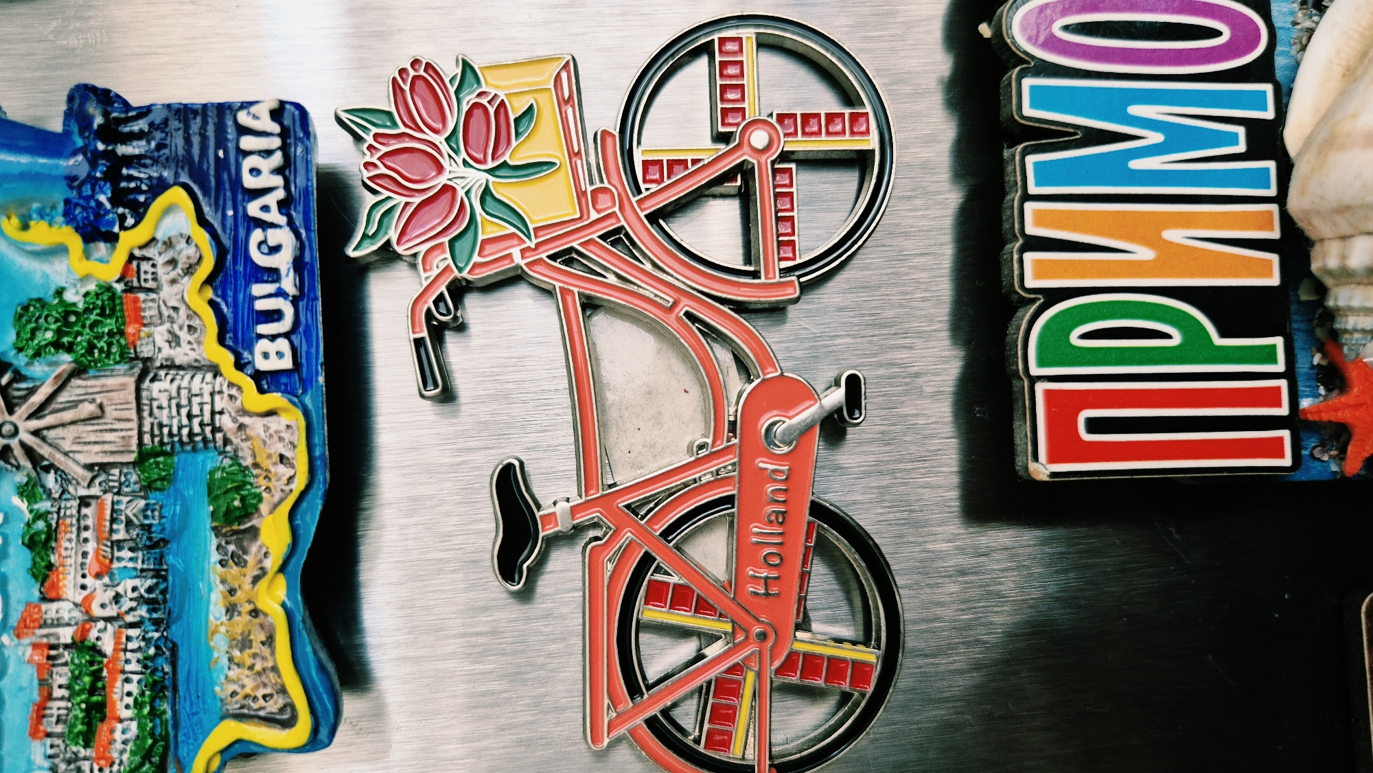 The image displays a collection of refrigerator magnets on a metallic surface. In the upper portion of the image, a magnet depicts a scene with buildings, a windmill, and the word "BULGARIA" written on it. Below this, a red bicycle magnet is prominently featured, with a basket of flowers on the handlebars and the word "Holland" written on the frame. Further down, a magnet with colorful letters spelling "ПРИМО" is visible, along with some seashells. The context of the city of Lovech, Bulgaria, is not visually evident in the provided image.