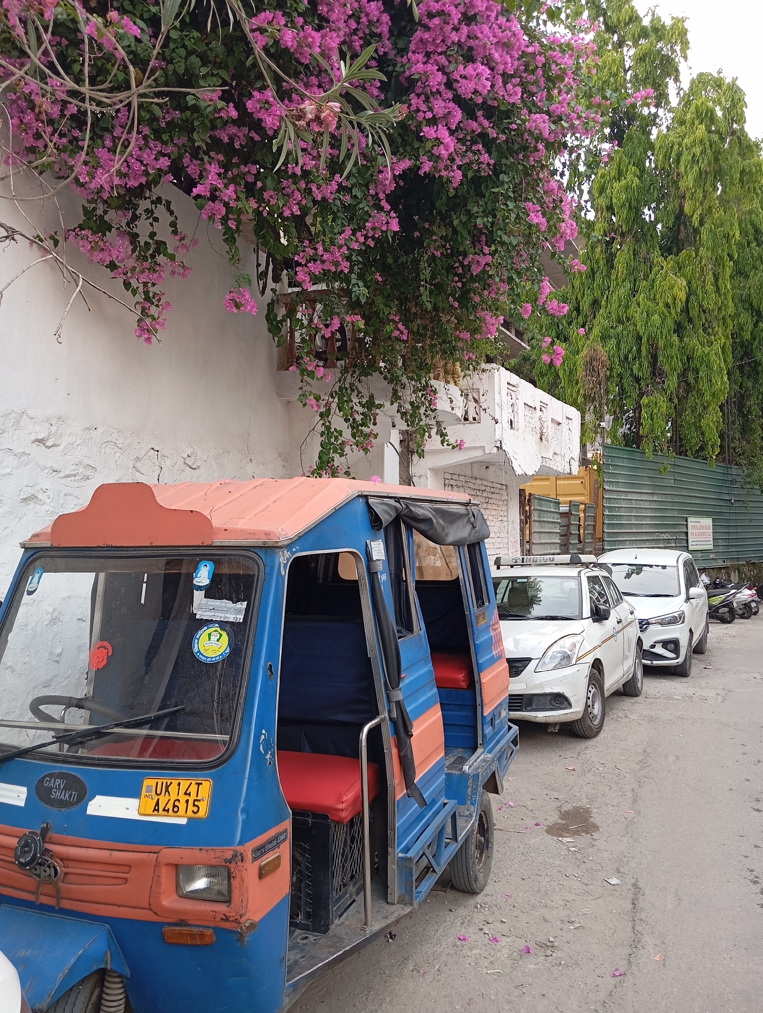 The image shows a street scene in the city of Unknown, Unknown. A blue and orange three-wheeled auto-rickshaw is parked on the side of the street. The rickshaw has a yellow license plate with the number "UK 14T A4615" and a sticker that reads "GARV SHAKTI." Behind the rickshaw, two white cars are parked in a row. In the background, a white building with bougainvillea vines growing on the wall is visible, along with tall green trees. A green fence is also visible in the background.