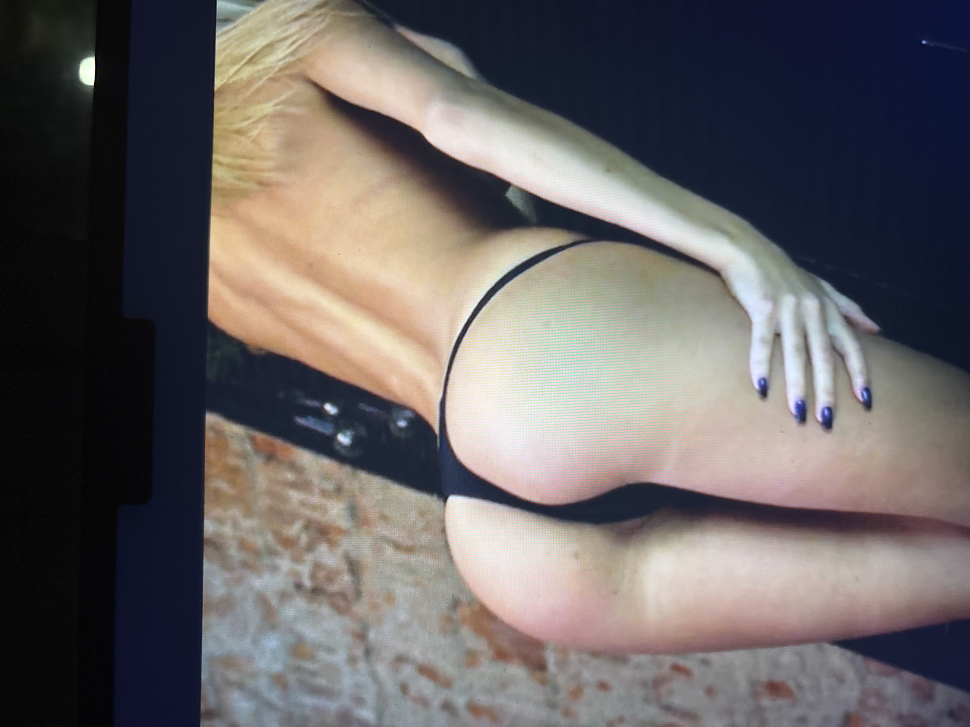 The image displays a partial view of a person, likely female, from the back, captured in an indoor setting in Sofia, Bulgaria. The subject is wearing a black thong bikini bottom, revealing her bare back and buttocks. Her right arm extends downwards from the upper frame, with her hand resting on her right upper thigh or hip. Her fingernails are painted a dark, possibly purple or dark blue, color. Light-colored hair, likely blonde, is visible on the upper left side of the frame.
The background is divided into two main areas: a dark, uniform surface occupies the upper right portion, while the lower half features a lighter, textured surface that resembles stone, brick, or patterned tile flooring. The person appears to be in a relaxed, possibly reclining or leaning, pose. The image itself is a photograph of a screen, indicated by the visible pixelation and a distinct bright, circular light reflection on the far left edge of the frame. No text is visible in the image.