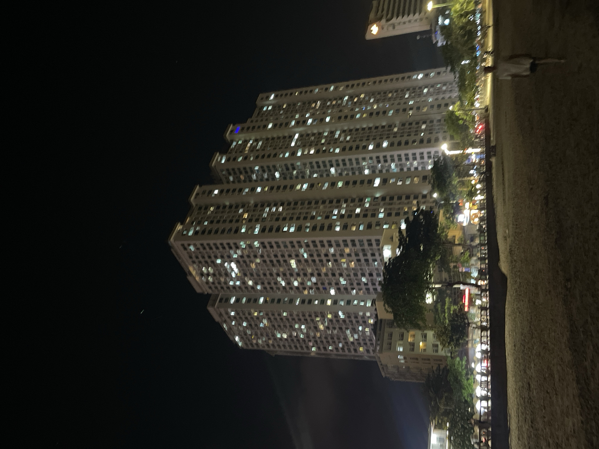 The image shows a tall, brightly lit building at night in Bắc Nha Trang, Vietnam. The building has a distinctive stepped design and numerous windows with lights on. To the right of the building is a pathway with trees and streetlights. A person is walking along the pathway. The sky is dark.