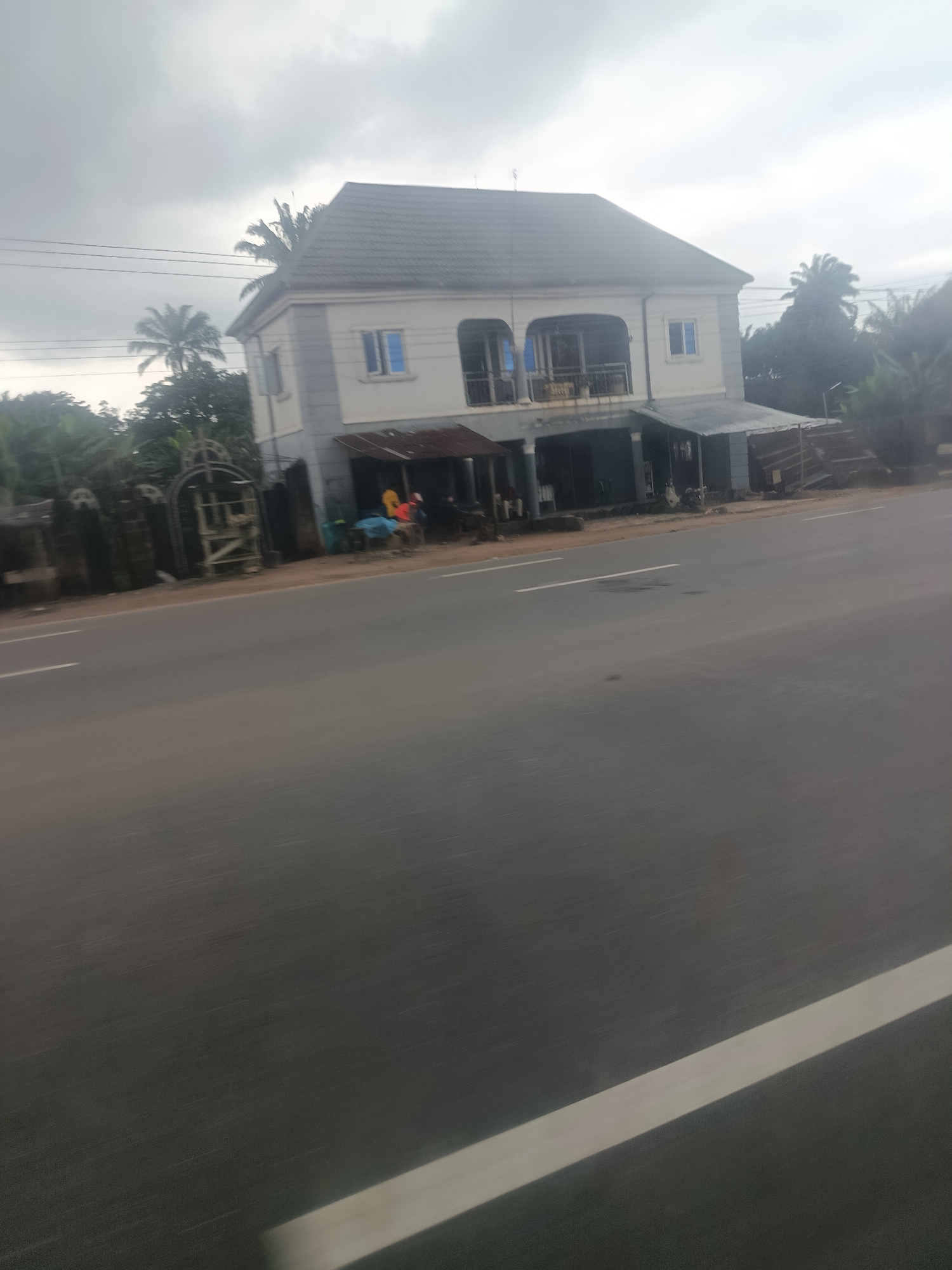 The image captures a street scene in Mgbidi, Nigeria, focusing on a two-story building situated adjacent to a road. The building has a white facade with gray accents and a dark-tiled roof. A balcony with railings is visible on the upper floor. The ground floor features a covered area that appears to house several small stalls or shops, indicated by corrugated metal roofing and visible goods. On the left side of the building, a dark, ornate gate or fence is present, suggesting an entrance to a property. In front of the building, a few figures, possibly vendors or customers, are present near the stalls. The road in the foreground is paved and marked with white lines. Palm trees and other lush vegetation are visible around the building and in the background, typical of the Nigerian landscape. The sky is overcast, contributing to a diffused, somewhat dim lighting condition.