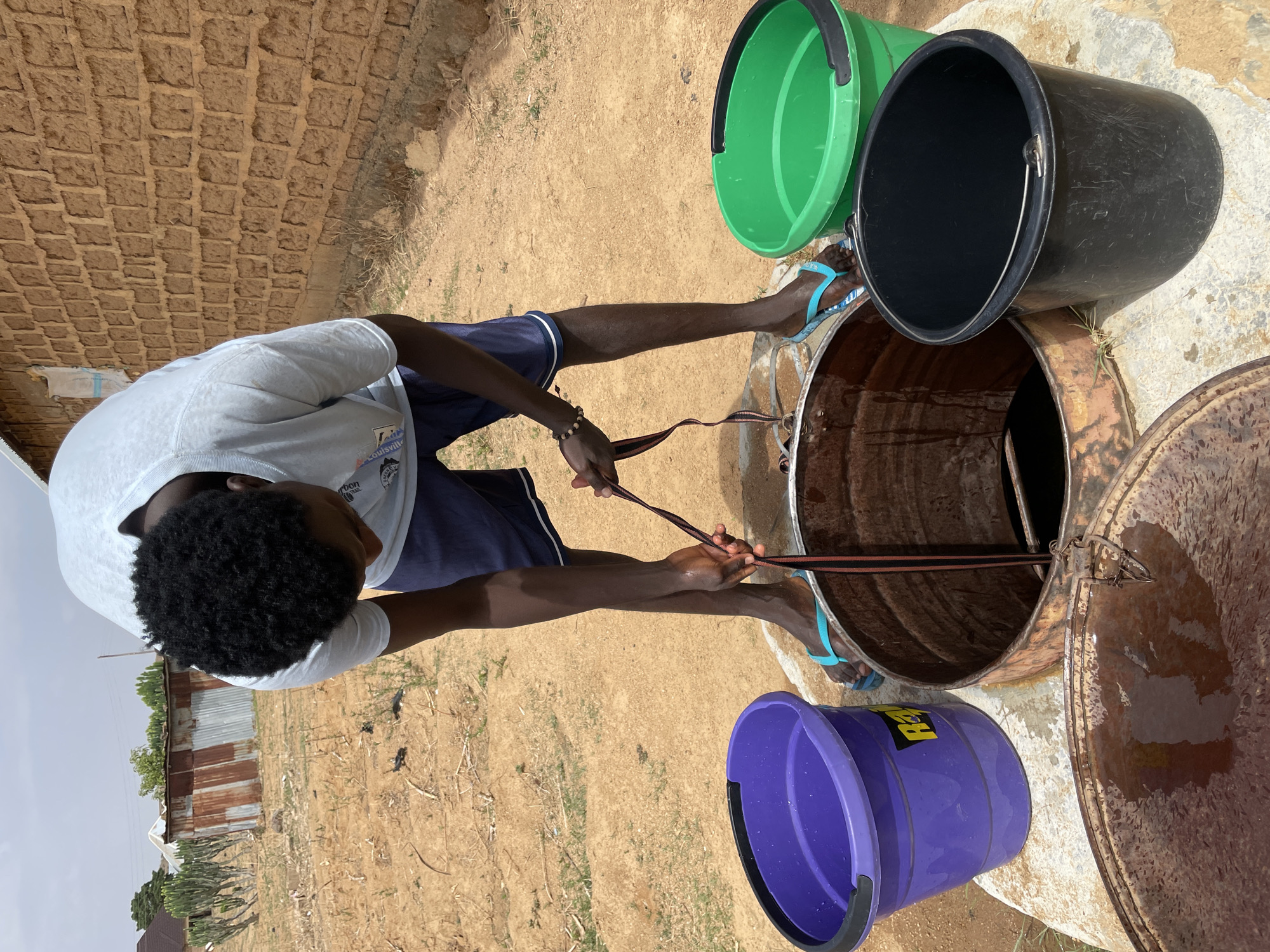 A man stands at a well in Jos, Nigeria. He is of African descent with dark curly hair, wearing a light-colored T-shirt, dark blue shorts, and blue flip-flops. He is holding a strap or rope, seemingly to draw water from the well. The well is constructed of rusted metal and is open with its lid ajar. Three buckets surround the well: one green, one black, and one purple. The background shows a dry, sandy landscape with buildings in the distance and a low, overcast sky. The area around the well is dirt, with some sparse vegetation. A brick or stone structure can be seen in the upper left corner of the frame.