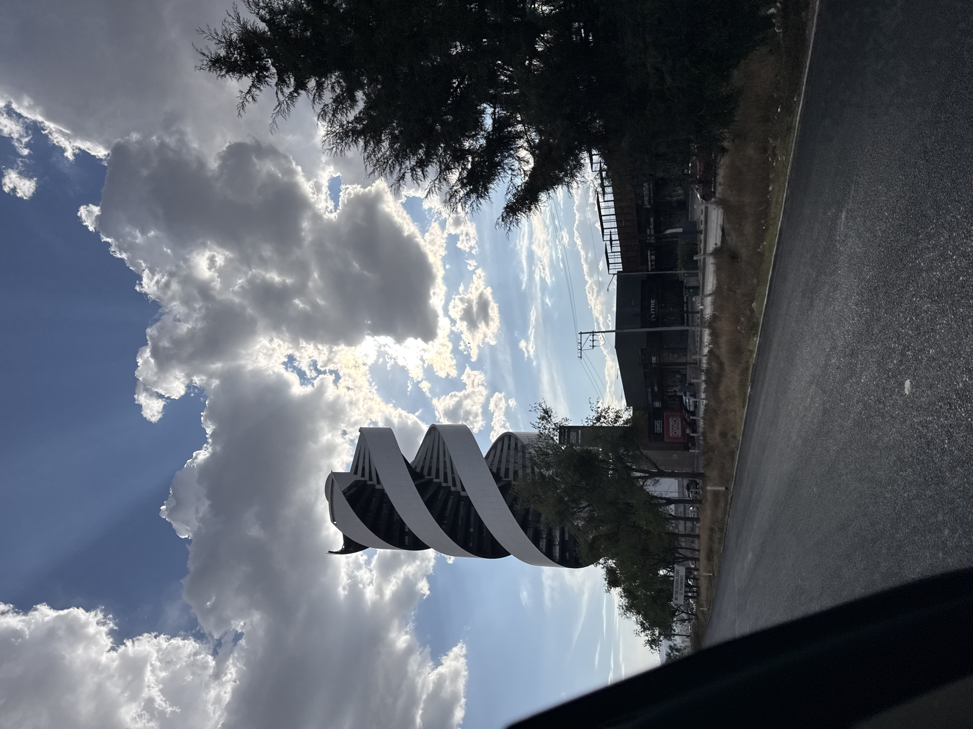 The image shows a building with a unique, helix-shaped design. The building's structure consists of a series of stacked, curved sections that spiral upwards. The building appears to be located in an urban area with other commercial establishments nearby. Trees and vegetation line the street. The sky is visible with a mix of blue sky and white, puffy clouds. The photograph was taken from a vehicle, with the edge of the dashboard visible in the foreground. The location is San Bernardino Tlaxcalancingo, Mexico.