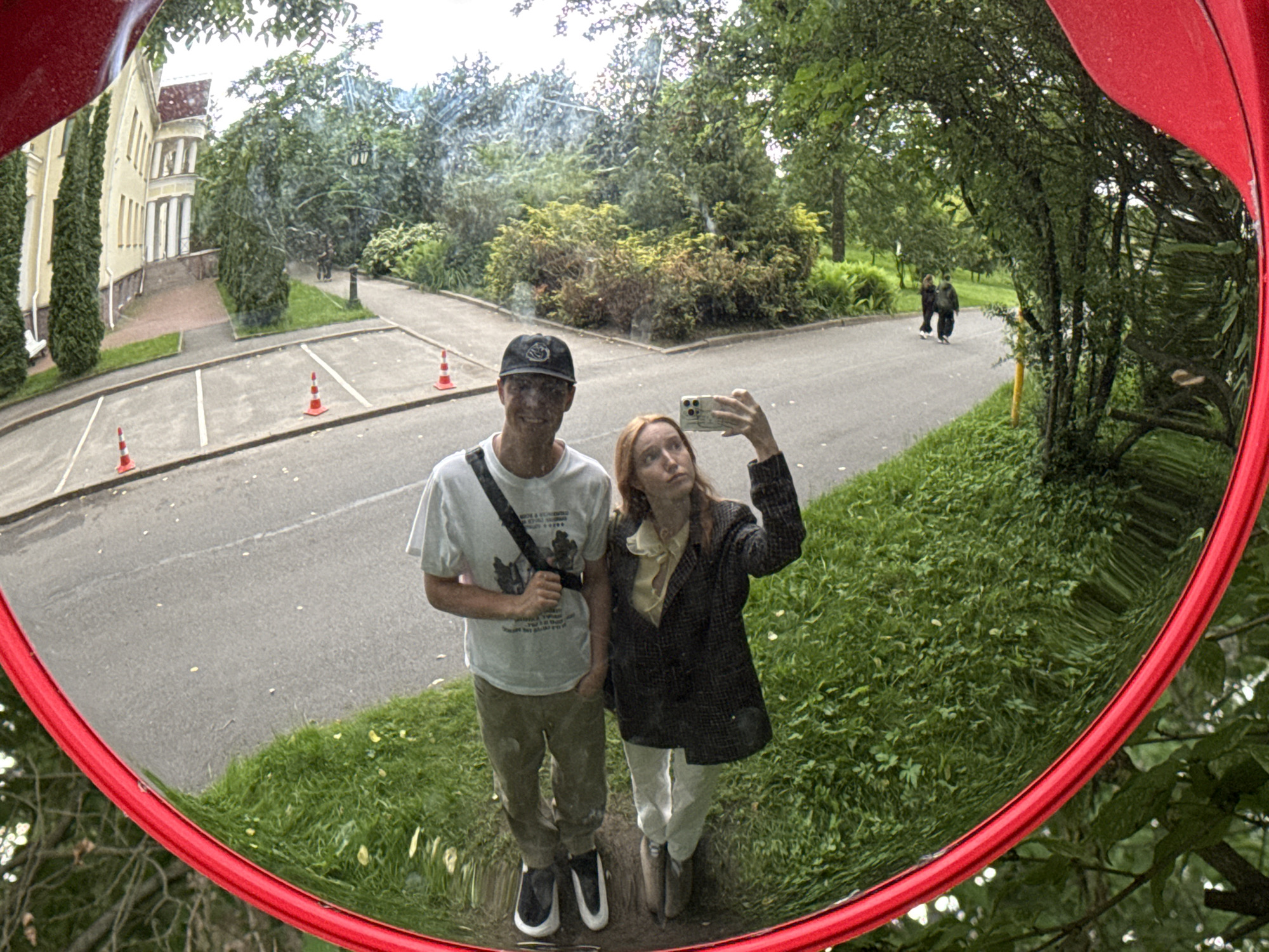 This is a fisheye lens reflection photograph taken in Saint Petersburg, Russia. The image shows a park-like setting with a building, trees, and a road. Two young adults, a male and a female, are taking a selfie. The male is wearing a white t-shirt, khaki pants, and a baseball cap. The female is wearing a patterned blazer over a yellow blouse and white pants. In the background, two other individuals are walking on the road. The reflection also shows orange traffic cones and parking spaces. The overall atmosphere is casual and relaxed.