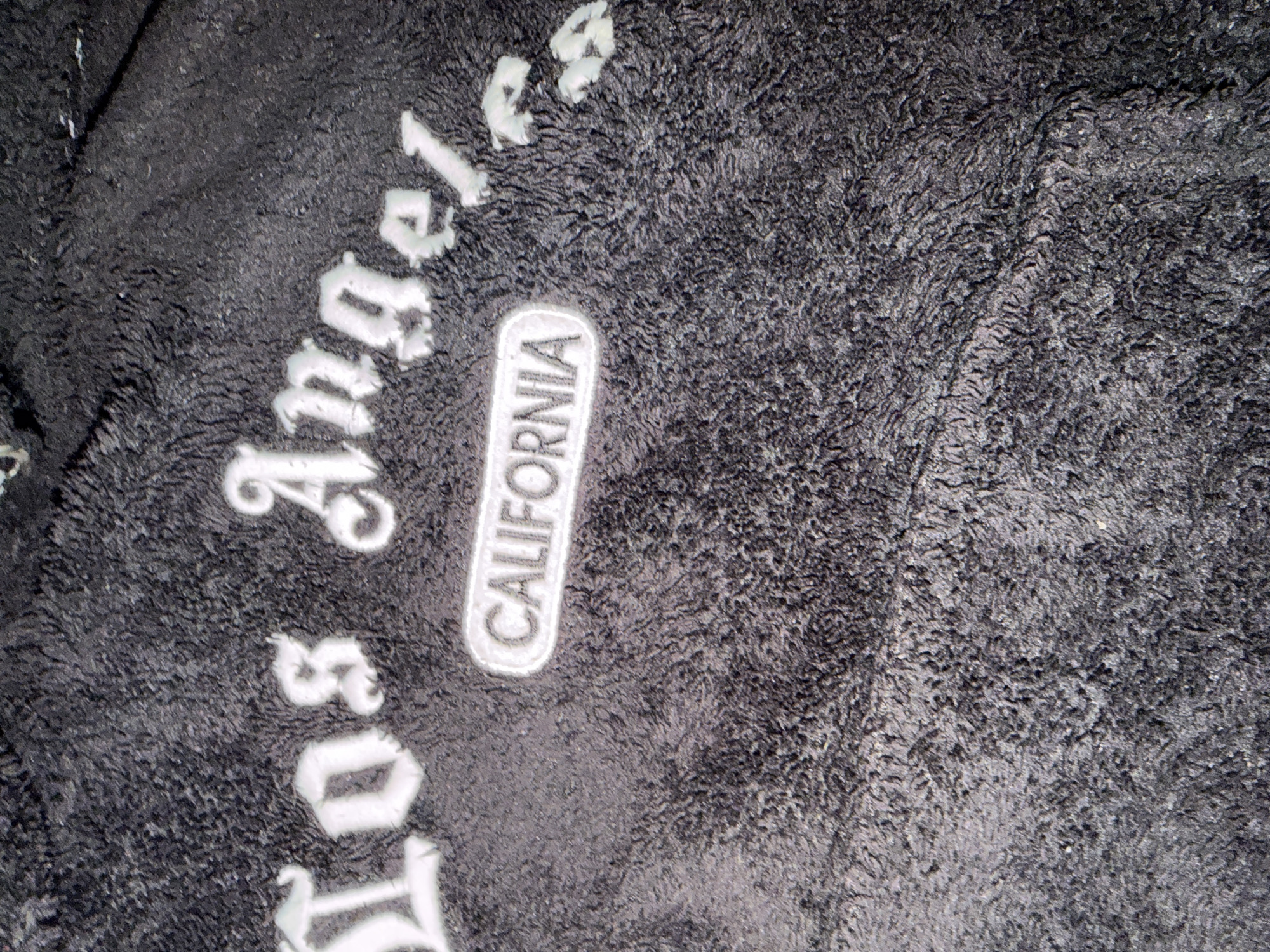 The image shows a close-up of a black, fuzzy fabric with white lettering that reads "Los Angeles" and "California." The lettering appears to be stitched onto the fabric. The background is out of focus, but appears to be more of the same fuzzy material. There is no indication of location in the city of Ivanski, Bulgaria, in the image.