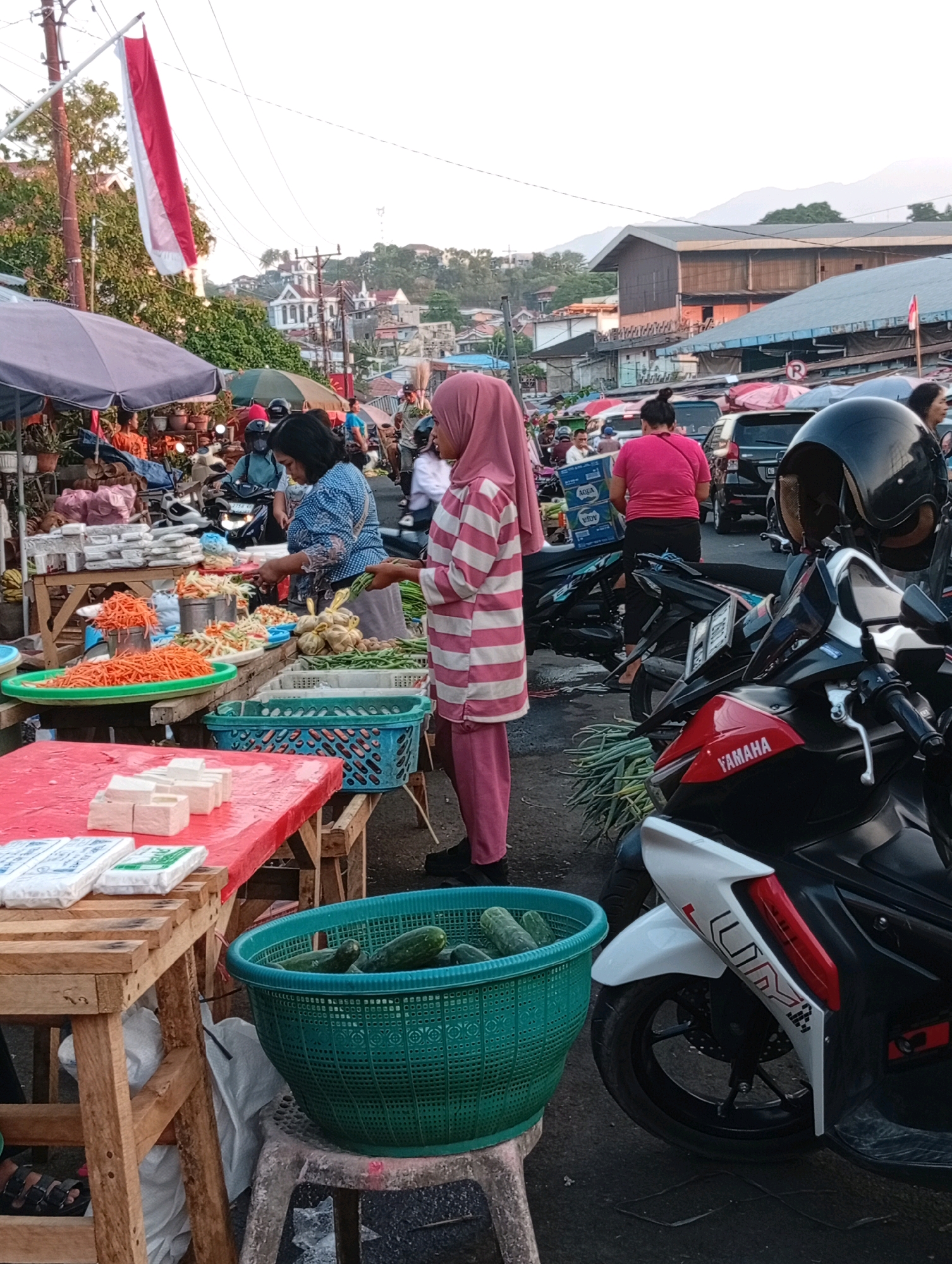 The image depicts a bustling street market in Manado, Indonesia. In the foreground, a wooden table displays stacks of white packaged goods, possibly tofu or soap. To the right, a green plastic basket overflows with fresh cucumbers. A scooter, partially visible, is parked on the right side of the frame, featuring "YAMAHA" branding on its fairing. A woman wearing a pink hijab and a striped pink and white outfit stands near the scooter, looking towards the left. Behind her, other market stalls are visible, with vendors and customers interacting amidst piles of produce and various goods. A prominent Indonesian flag, red and white, is flown from a pole on the left side of the image, adding a patriotic element to the scene. The background reveals a hilly cityscape with buildings and trees, suggesting a vibrant and active urban environment.