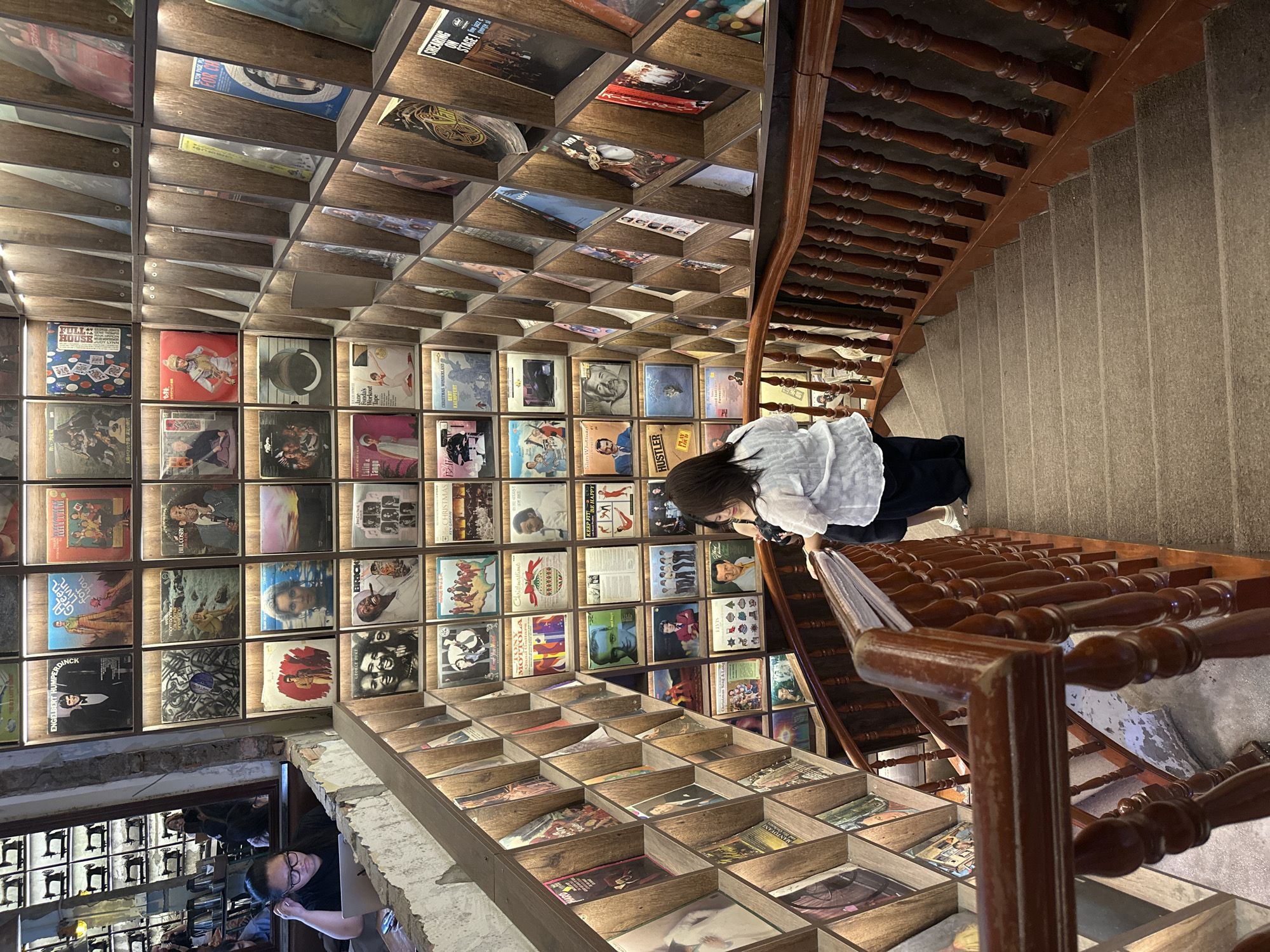 The image captures the interior of a record store in the city of Thủ Đức, Vietnam. A vast display of vinyl record albums covers the walls and ceiling, arranged in a grid-like pattern, creating a visually captivating atmosphere. A staircase with dark wooden railings and light gray steps ascends through the store. A woman in a white blouse and dark pants stands on the stairs, browsing the records. She appears to be examining the albums displayed on a shelf along the staircase. Further down, another individual, a woman wearing glasses, sits in what appears to be a seating area, partially visible through a reflection in a glass panel. The store is well-lit, highlighting the textures and colors of the records and interior architecture.