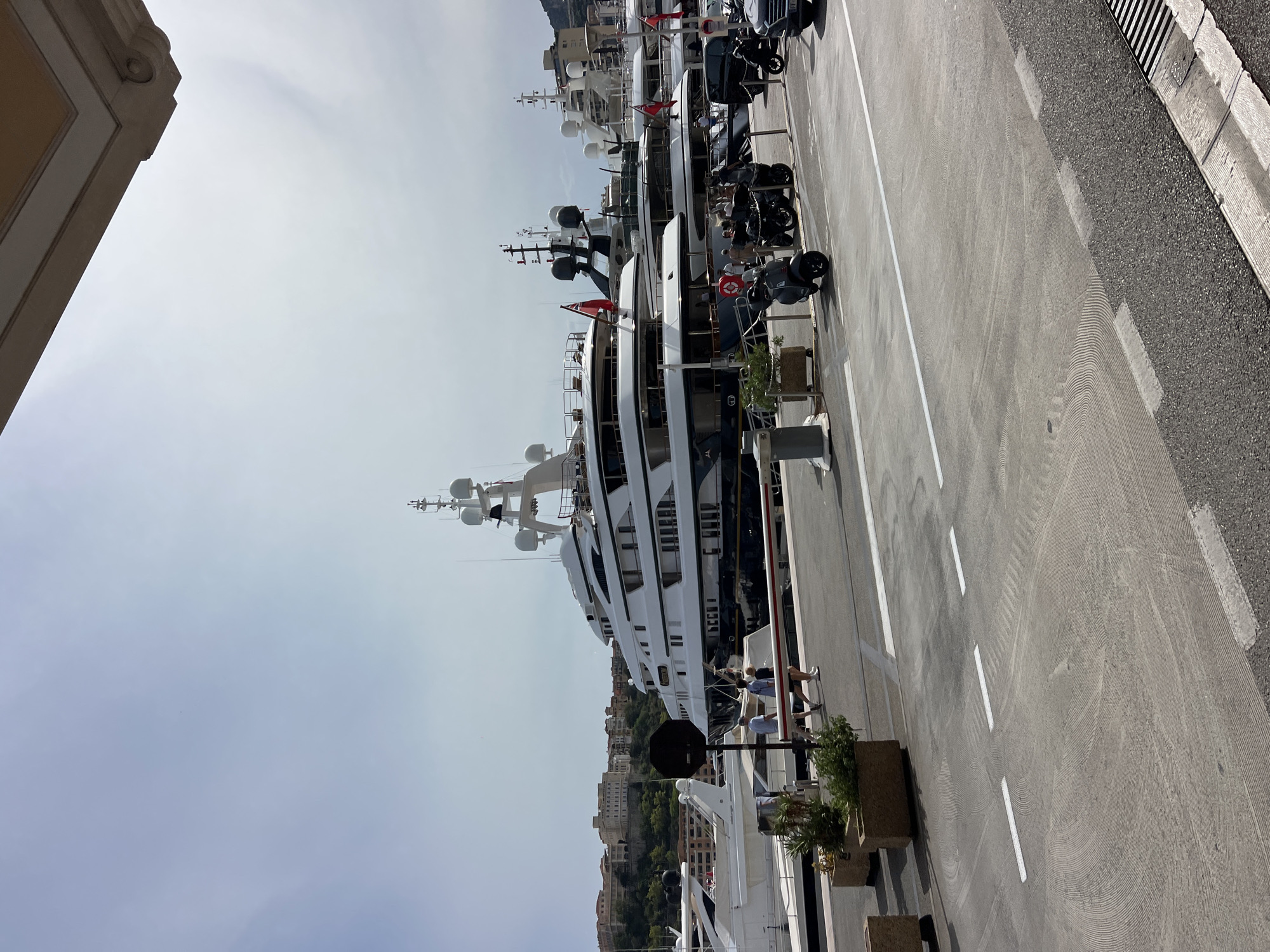 The image shows a sunny day in a marina in Monaco, Monaco. Several large yachts are docked, with one particularly prominent white yacht featuring multiple decks and a dark hull. In the foreground, a paved area with white lane markings is visible, with several scooters and a car parked on the side. A few people are walking along the dock, and potted plants are placed along the waterfront. In the background, a hillside with buildings and vegetation slopes upwards. The sky is partly cloudy.