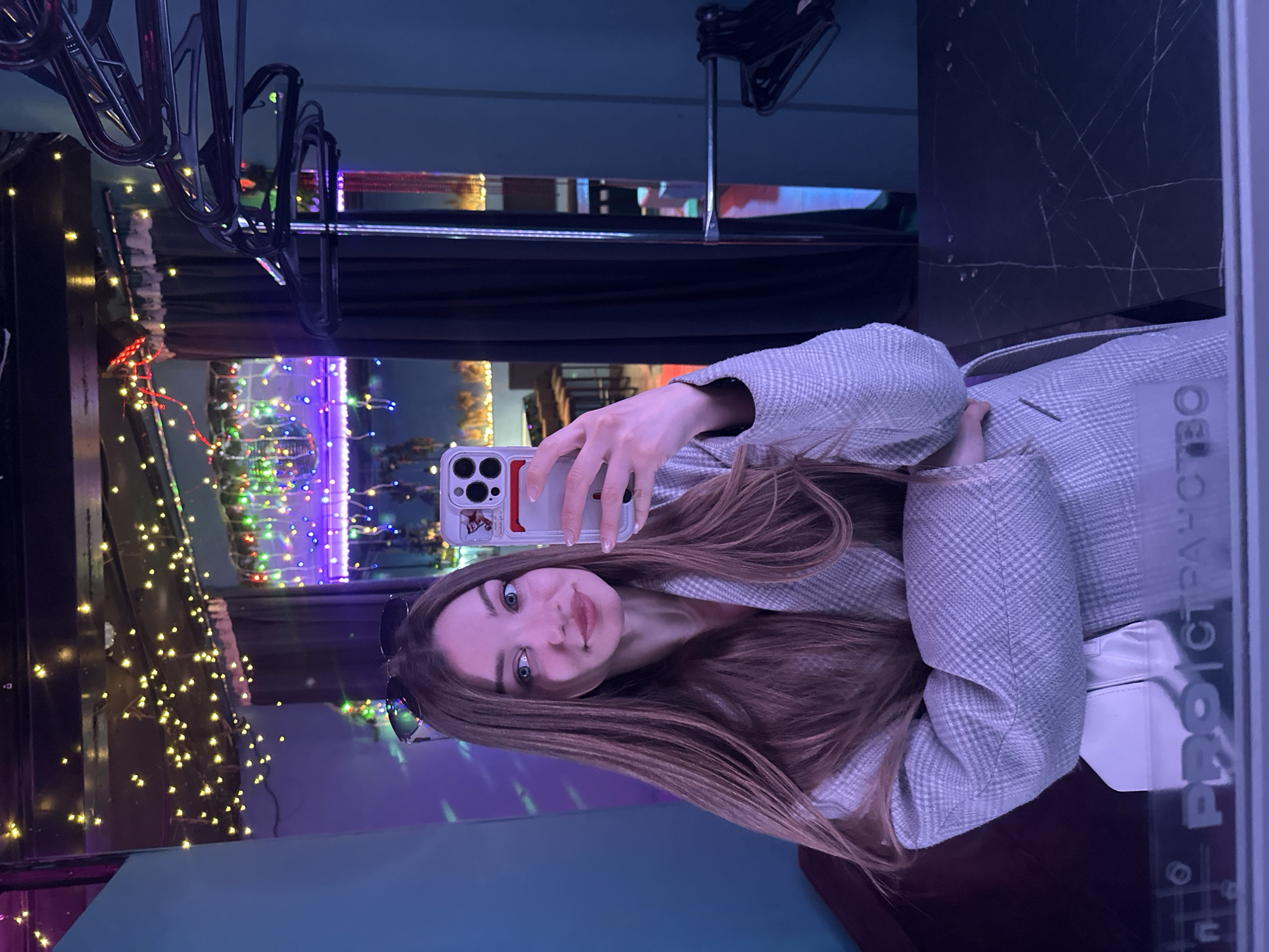 A young woman is taking a selfie in front of a mirror, holding a phone with a decorative case. She has long brown hair and is wearing a light gray plaid jacket. Sunglasses are visible on her head. The background features a wall with hanging clothes and a lighted display. The reflection in the mirror suggests the location is in Kazan, Russia.