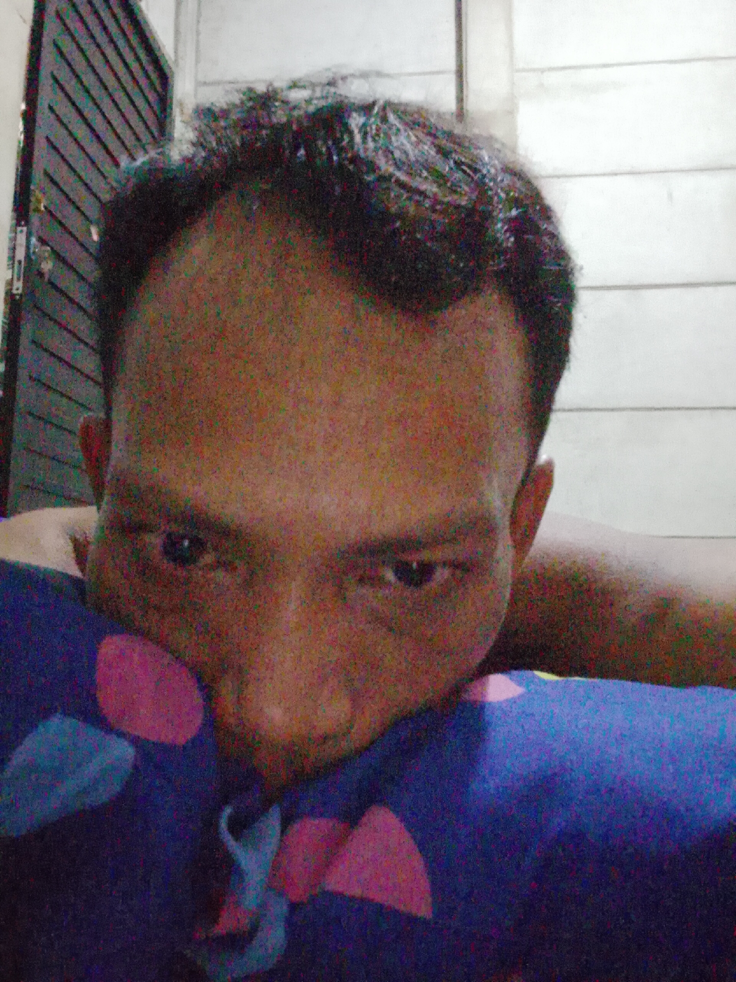 A close-up selfie of a man is seen against a wall that appears to be lined with planks, or a door, and plain wall. He is lying on a pillow with pink and blue designs. The man's face is prominent, with his eyes looking directly at the camera. The man appears to be of Southeast Asian descent. The location is stated as Bukit Ketok, Indonesia.