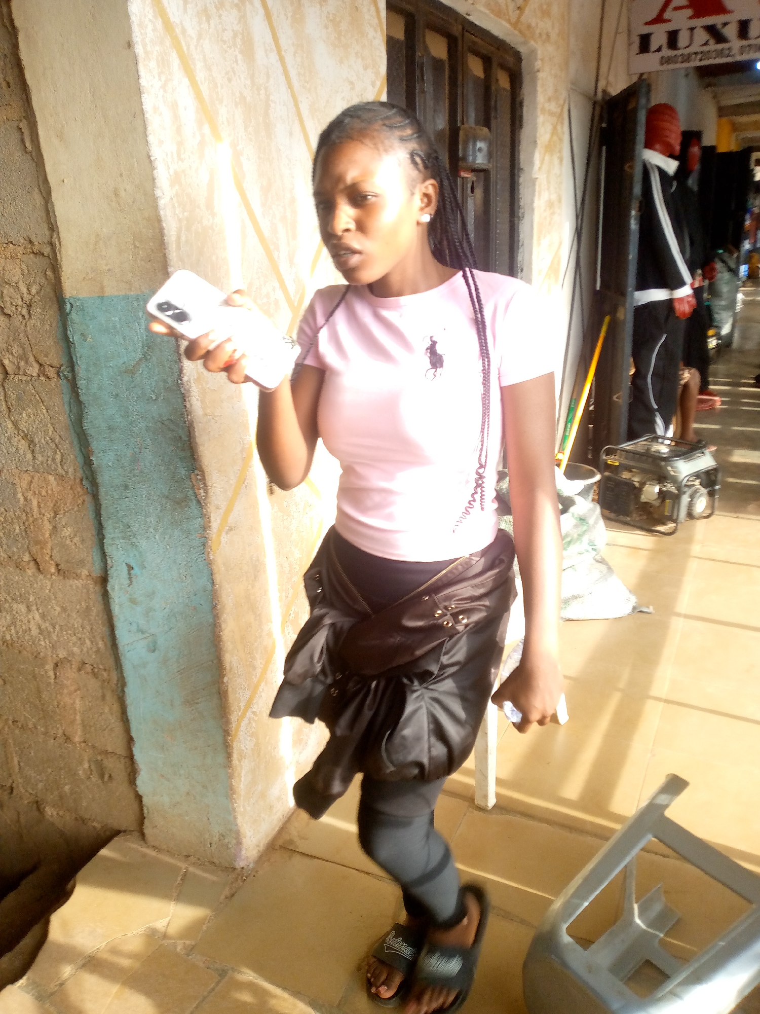 A young woman stands in a tiled hallway in Bukuru, Nigeria. She is holding a white smartphone to her left ear with her right hand and another smartphone in her left hand. She is wearing a light pink t-shirt with a black equestrian logo on the left chest, black leggings, and black slide sandals. A black fanny pack is tied around her waist. Her hair is braided and pulled back. To her left is a weathered wall with a blue-painted section. Behind her to the right, is a doorway leading into a store called LUXU with a mannequin in a black suit and red helmet. A generator sits in the right background, along with other people in the store. A broken chair is to her lower right. The floor is bright, presumably due to natural sunlight coming into the hallway.