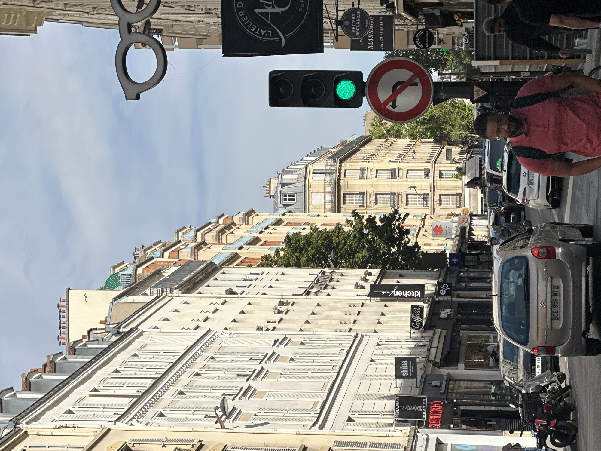 This is a street-level, medium-shot photograph taken in Paris, France, likely on a sunny day with some clouds. The perspective is from the street, looking down a bustling urban street lined with multi-story Haussmannian-style buildings. These buildings feature cream-colored facades, numerous windows with white shutters, and grey mansard roofs.
In the foreground, a gold-colored Citroen car with the license plate "CE-959-NT" is prominent, partially obscuring the view of the street ahead. To the left of the car, several motorcycles are parked. Further down the street, traffic is visible, including other cars and a white taxi.
A man wearing a red polo shirt and a backpack is walking towards the camera, looking down. Several other pedestrians are visible in the background, as well as various shops and businesses lining the street. Signs visible include "RESTAURANT JAPON," "Shiva," "Coiffure," "kitchenlab," and a large sign with an emblem resembling glasses.
Traffic lights and street signs are also present. A traffic light with a green light illuminated is visible, along with a circular red and white sign indicating "no left turn." The sky above is a pale blue with scattered clouds. The overall impression is one of a vibrant, active Parisian street scene.