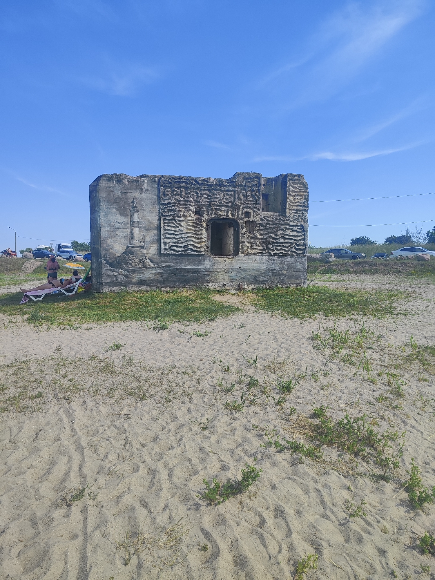 In Rovnoe, Russia, a concrete structure with a lighthouse mural and wave-like carvings sits on a sandy beach under a clear blue sky. In front of the structure, two individuals are relaxing on lounge chairs under a beach umbrella. The background shows a road with several parked vehicles and vegetation.