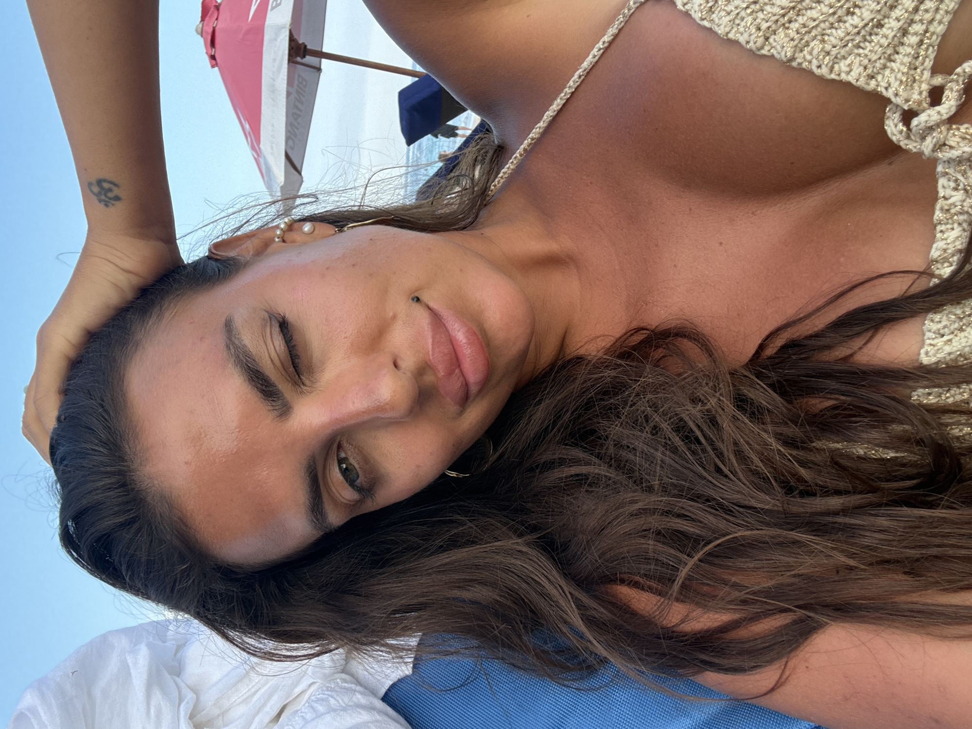 A close-up, selfie-style photo shows a woman with long, dark brown hair, a tanned complexion, and green eyes. She is winking, with one eye closed and the other open. Her left hand is raised to the back of her head, and she is wearing a cream-colored, crocheted bikini top. A small black mole is visible on her upper lip, and she has a tattoo on her left forearm. In the background, a pink beach umbrella is partially visible, along with lounge chairs, a body of water, and a clear blue sky. The setting appears to be a beach in Kuta Selatan, Indonesia.