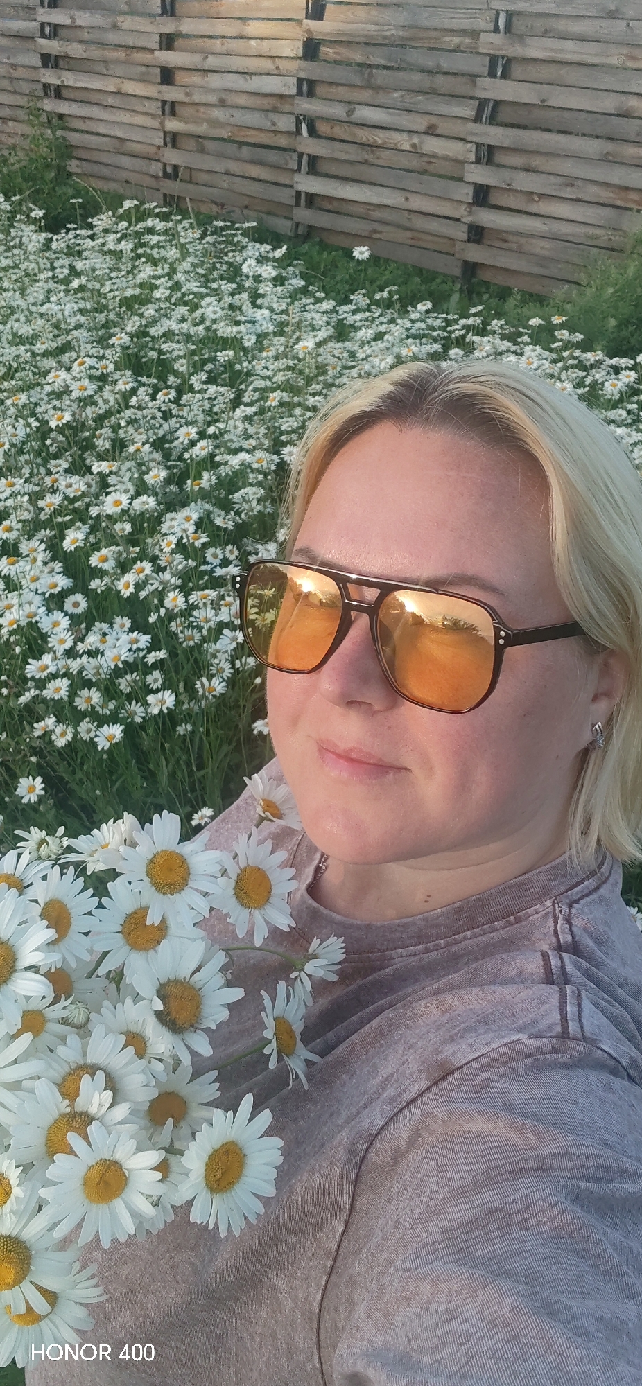 A blonde woman with sunglasses and a bouquet of daisies poses for a selfie in a field of daisies. She is wearing a taupe t-shirt. The sunglasses have orange lenses and reflect the sky. Behind her is a wooden fence and more daisies. The scene suggests it is daytime and likely warm weather, given the setting and attire. The image appears to be taken outdoors in a rural or natural environment. The overall mood is relaxed and cheerful. "HONOR 400" is visible in the bottom left corner of the image.