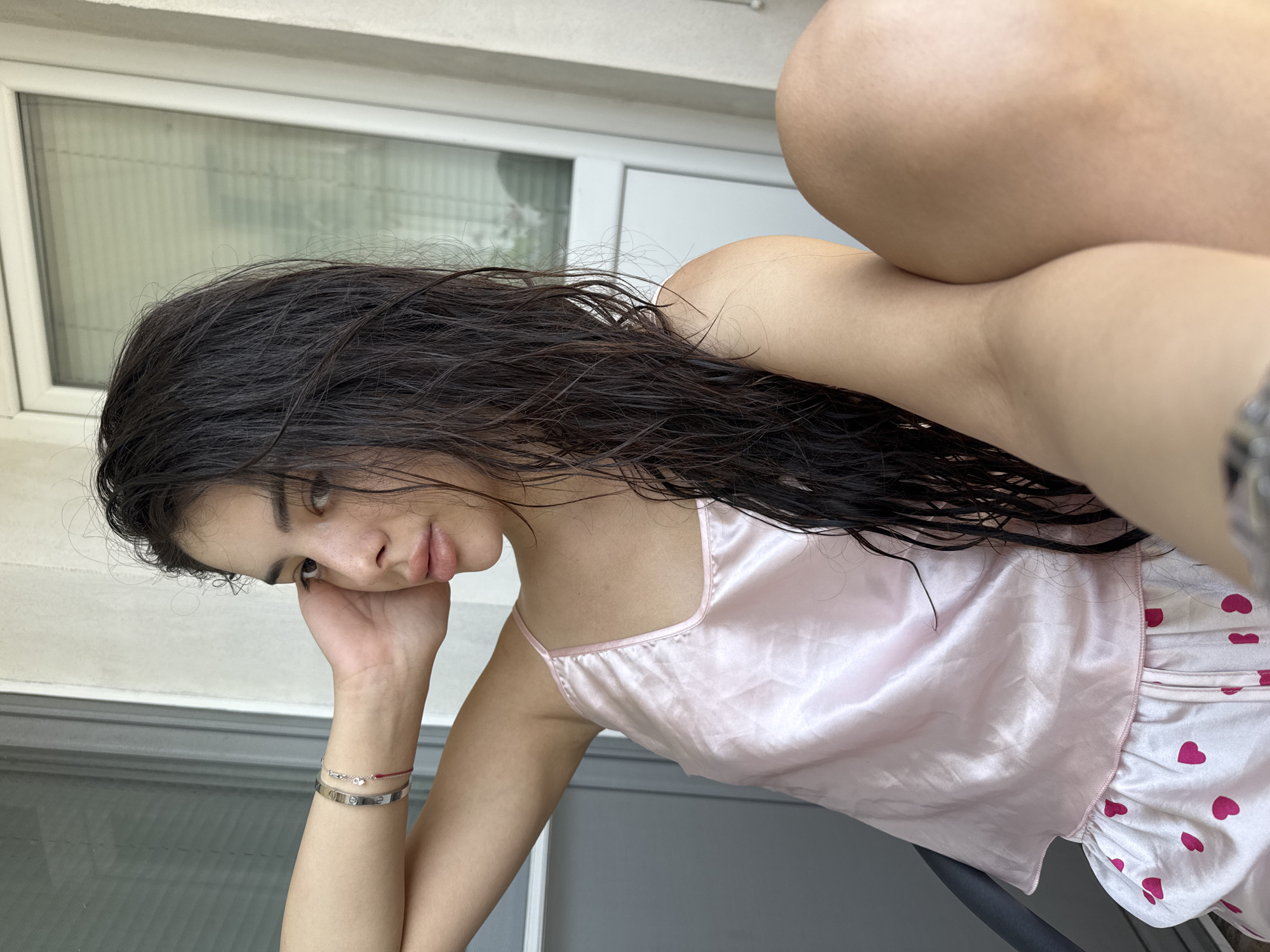 The image displays a young woman seated on a balcony or terrace in Plovdiv, Bulgaria, captured in an eye-level, slightly upward-angled perspective. She has long, wet, dark hair draped over her shoulders and chest. Her left arm is raised, with her hand resting against her temple. On her left wrist, she wears a silver-toned bracelet with screw motifs (resembling a Cartier Love bracelet), a thinner silver-toned bracelet with a small charm, and a delicate red string bracelet. She has a fair complexion with visible freckles on her face and full lips. She is dressed in a light pink satin camisole with thin straps and light-colored shorts adorned with a pattern of scattered pink hearts. The background features light-colored walls and two window or door structures. On the left, a dark-framed sliding glass door or window with visible horizontal blinds can be seen. On the right, a white-framed window or door, also with interior blinds, is present. A dark, curved object, possibly part of a chair or railing, is visible in the lower-left foreground. Her right leg is partially visible and bent in the foreground, indicating a relaxed, seated posture.