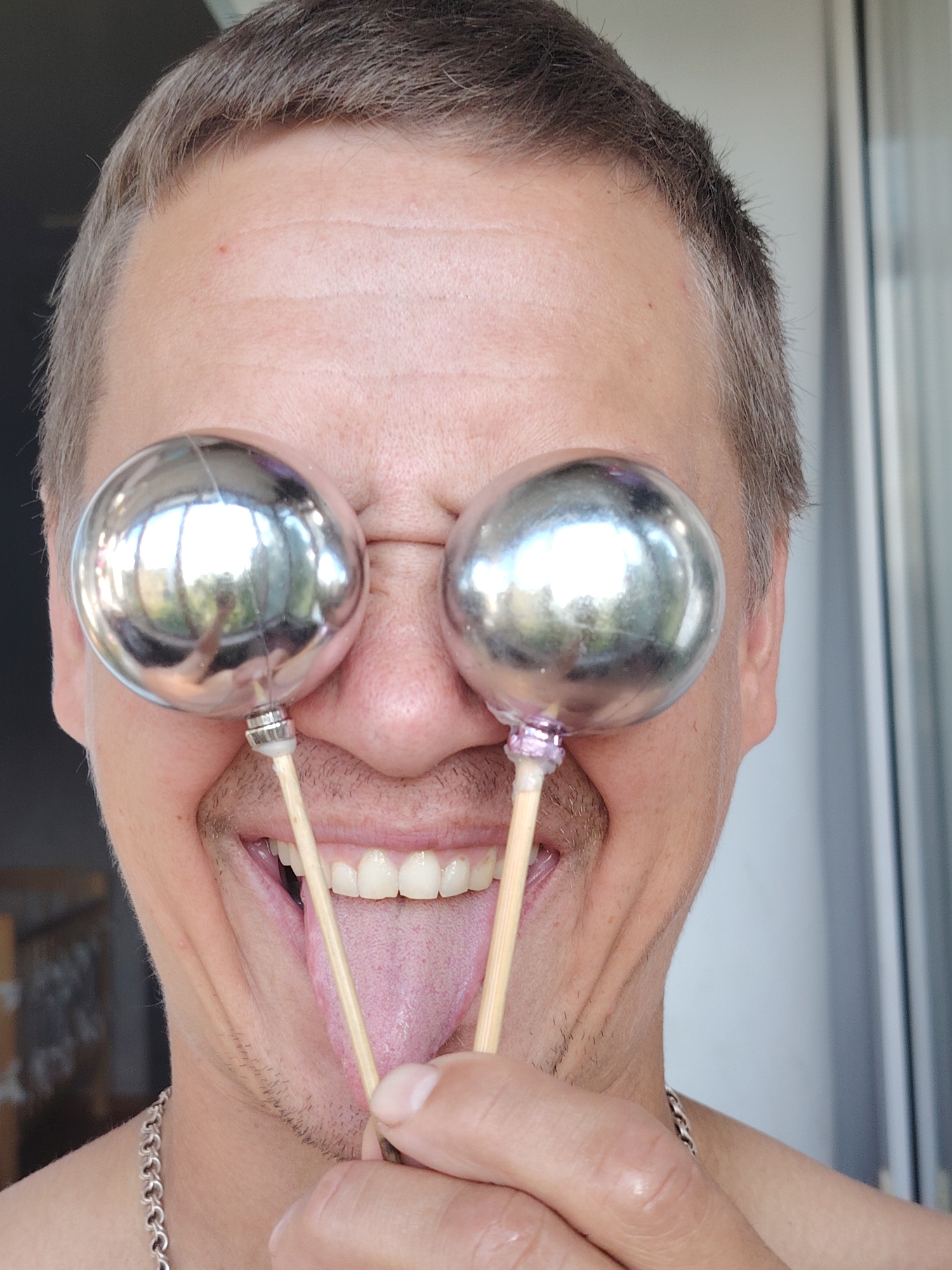 The image shows a close-up of a person's face, with their mouth open and tongue sticking out. Two spherical, metallic objects are held in front of the person's eyes by wooden sticks. The objects appear to be decorative balls. The person has short, light brown hair and is wearing a silver chain around their neck. The background suggests an outdoor setting with a building or wall visible to the right. The image does not contain any information that would allow for identification of its location in the city of Chaikovsky, Russia.