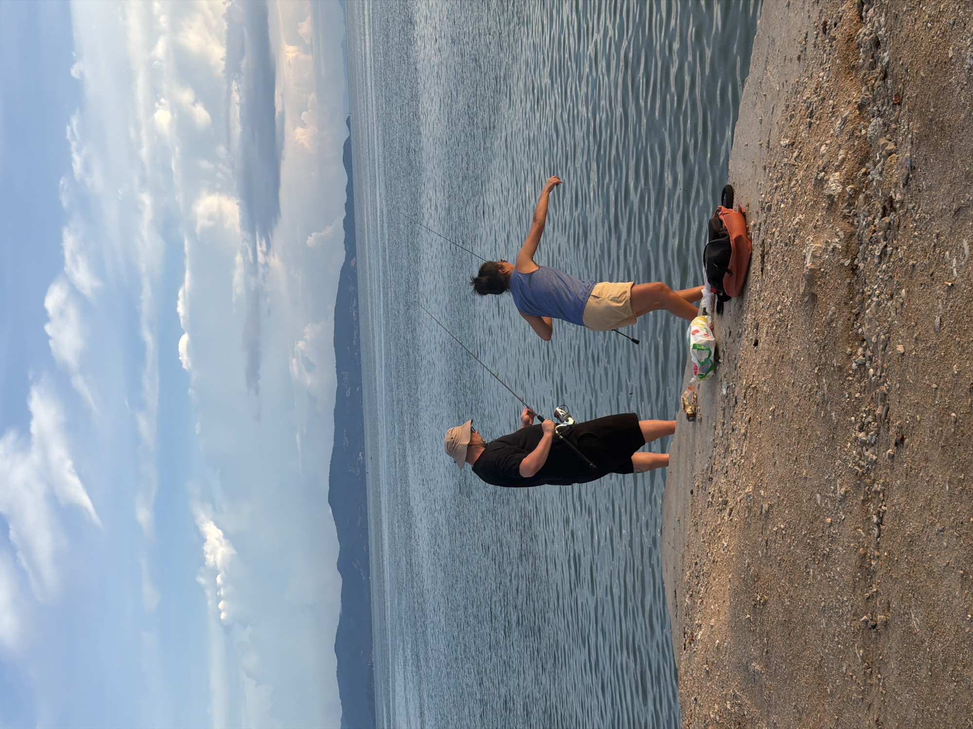 Two people are fishing on the edge of a concrete structure overlooking a body of water. The person on the left is a man wearing a black t-shirt, black shorts, and a light-colored hat. He is holding a fishing rod with a reel. The person on the right is a woman wearing a blue tank top and tan shorts. She is also holding a fishing rod, and appears to be in the process of casting or reeling in. In the background, there is a hazy view of mountains and the sea, under a sky with scattered clouds. On the ground near the people are two bags and a plastic bag. The location is identified as ตำบลเกาะพะงัน, Thailand.