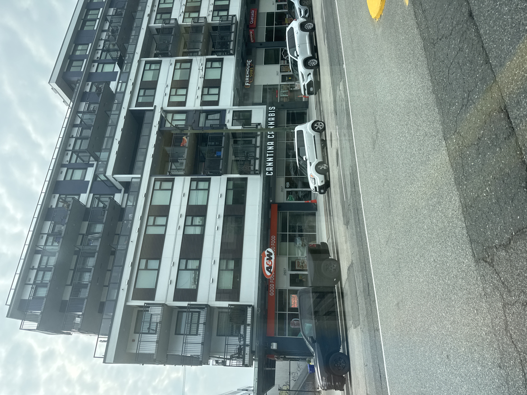 An exterior view shows a modern, multi-story building with a mix of residential apartments and commercial establishments on the ground floor. The building features white and dark blue facades, with multiple balconies with black railings. Below the apartments, the commercial storefronts are visible, including an "A&W" restaurant and "Cannitina Cannabis." Other businesses like "Firehouse Subs" and "Quesada" are also present. Several vehicles are parked along the street in front of the businesses, including a black pickup truck and white SUVs. The scene appears to be in a city environment, with a cloudy sky overhead. The location is identified as Vancouver, Canada.