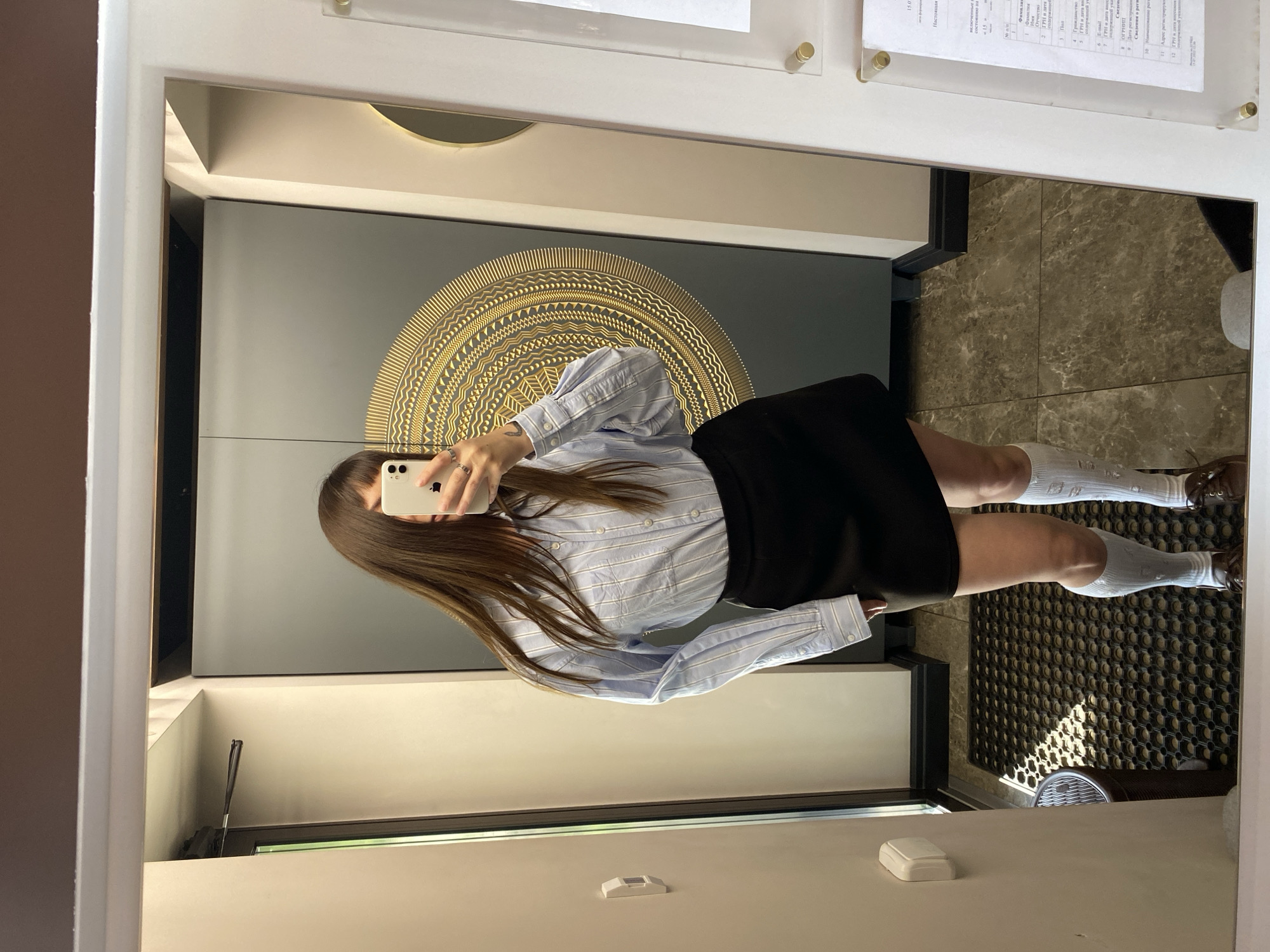 A young woman with long brown hair, wearing a light blue and white striped button-down shirt, a black skirt, white knee-high socks with distress details, and brown loafers, stands in front of a large mirror. She is holding a white smartphone in her right hand, taking a mirror selfie. Her face is obscured by her hair and the phone. The background shows a gray door with a large, circular gold decorative pattern. To the left of the door, a portion of a white wall with a light switch is visible, and to the right, a reflective surface, possibly another mirror or glass panel, displays some text. The floor is tiled with a marble pattern, and a black mat is positioned in front of the door. The scene appears to be an interior hallway or entryway, with natural light entering from the left side. The location is identified as Votkinsk, Russia.