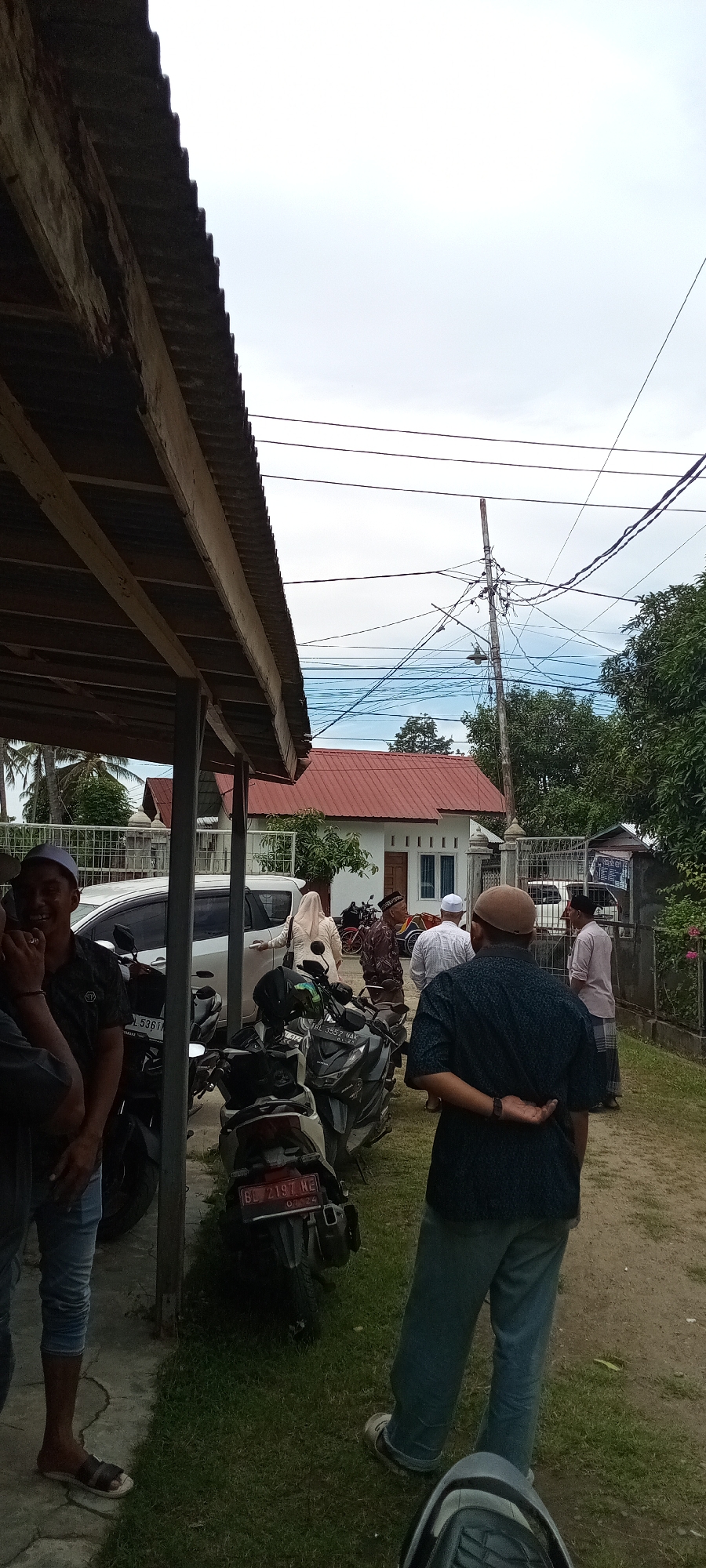 The image depicts an outdoor gathering in what appears to be a residential or communal area in Lhokseumawe, Aceh Province, Indonesia, as indicated by visible "BL" region license plates on two motorcycles ("BL 5361" and "BL 2197 HE"). The time of day is characterized by an overcast sky, resulting in diffuse lighting and minimal strong shadows.
In the foreground, a dark corrugated metal roof, supported by rough wooden beams, extends from the left, partially shading the scene. Beneath this shelter, several people are present. On the far left, a man wearing a white peci (prayer cap), a dark polo shirt, blue jeans, and sandals is seen smiling towards the left, with part of his face obscured by another person's arm. To his right, another man, viewed from the back, wears a brown cap, a dark blue patterned short-sleeved shirt, and light blue trousers, with his hands clasped behind his back.
Multiple motorcycles are parked on the unpaved ground, which is partially covered with grass. Directly behind the man facing away, a silver motorcycle with license plate "BL 2197 HE" (dated 03.24) is visible. Further left and behind, another motorcycle with license plate "BL 5361" is parked.
In the mid-ground, a single-story white house with a reddish-orange tiled roof stands centrally, featuring a brown wooden front door and three small vertical windows to its right. A white metal fence and gate define the property line in front of the house. To the right of the white house, a wooden utility pole is prominent, festooned with numerous black electrical wires and a single attached light fixture, extending upwards against the cloudy sky.
Several individuals are gathered around the white house and a parked white multi-purpose vehicle (MPV). A woman in a light-colored headscarf and dress is observed opening the rear passenger door of the white MPV. Other men, many wearing peci or kopiah, are conversing or standing in proximity to the house and vehicles. On the far right, a man in a light pink shirt and a sarong is visible. Behind the people on the right, a red three-wheeled vehicle is partially discernible.
The background features lush green foliage, including several trees and palm trees on the left, indicative of a tropical environment. The ground throughout the visible area is a mix of packed dirt and patchy grass.