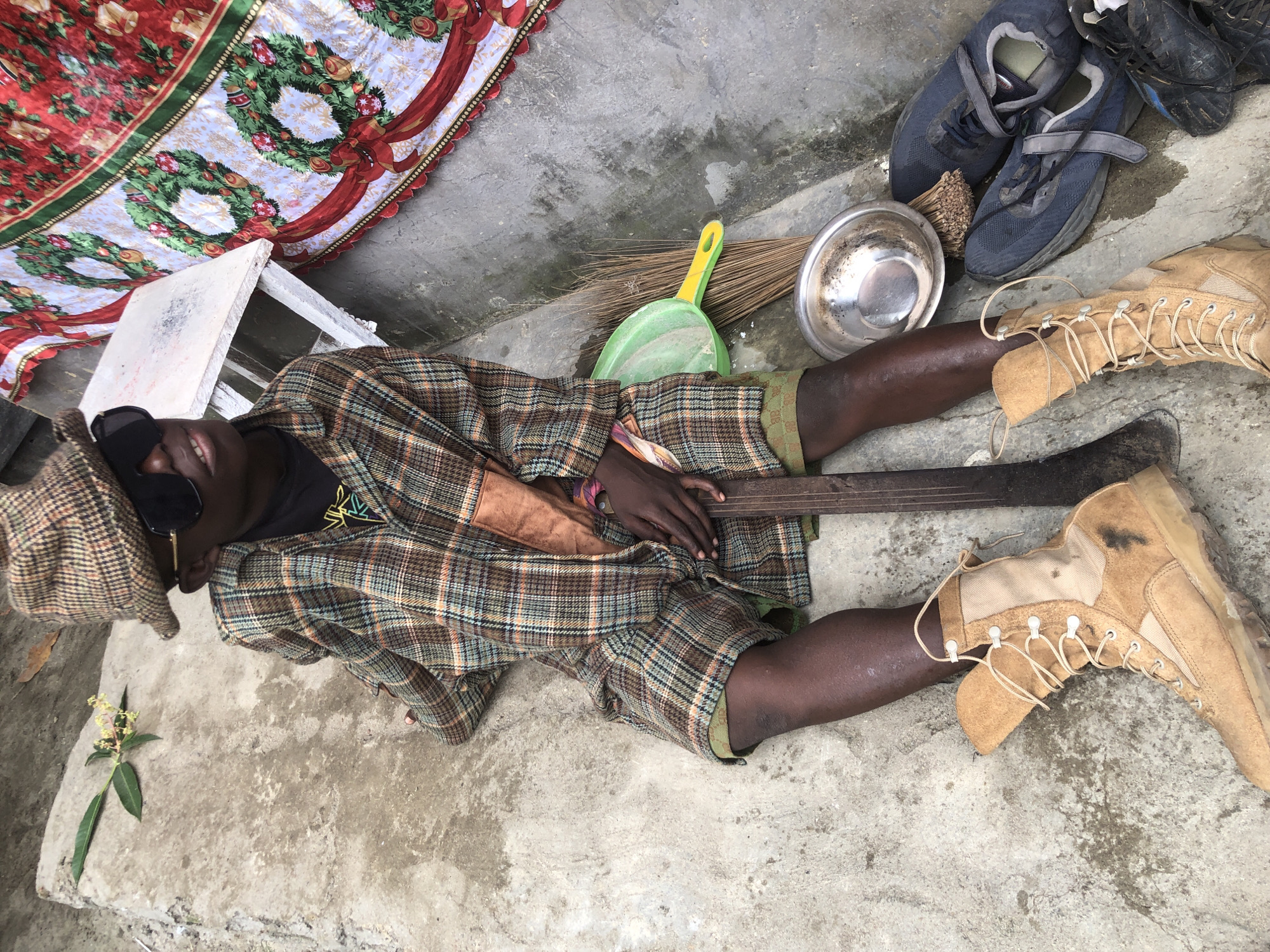 A dark-skinned individual, wearing a plaid hat, large dark sunglasses, and a plaid shirt and shorts, sits on a concrete surface. The individual is holding a wooden object. To the right of the individual are cleaning supplies, including a broom and dustpan, a metal bowl, and a pair of blue sneakers. A colorful, festive cloth with wreath motifs is draped in the background. The scene appears to be outdoors in Ado, Nigeria.