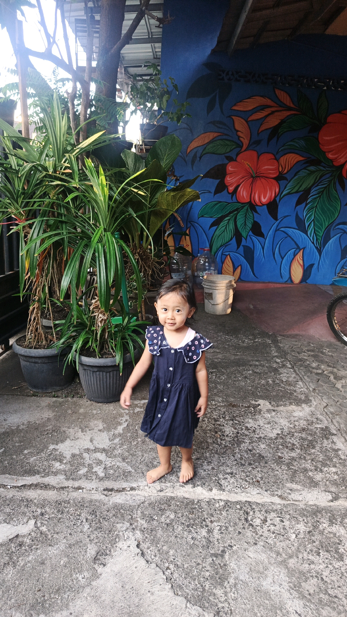 A young girl wearing a dark blue dress stands on a concrete ground in front of a vibrantly painted mural on a blue wall. The mural depicts large red hibiscus flowers with green and orange leaves. To the left of the girl are several potted plants with lush green foliage, including one very tall plant in a black pot. In the background, behind the plants, are two clear plastic water bottles and a light-colored bucket. The environment appears to be an outdoor space, possibly a courtyard or alleyway, with overhead roofing and natural light filtering through trees. The ground is rough and uneven concrete. The girl is facing the camera with a slight smile and appears to be standing barefoot. The scene is located in the city of Surakarta, Indonesia.
