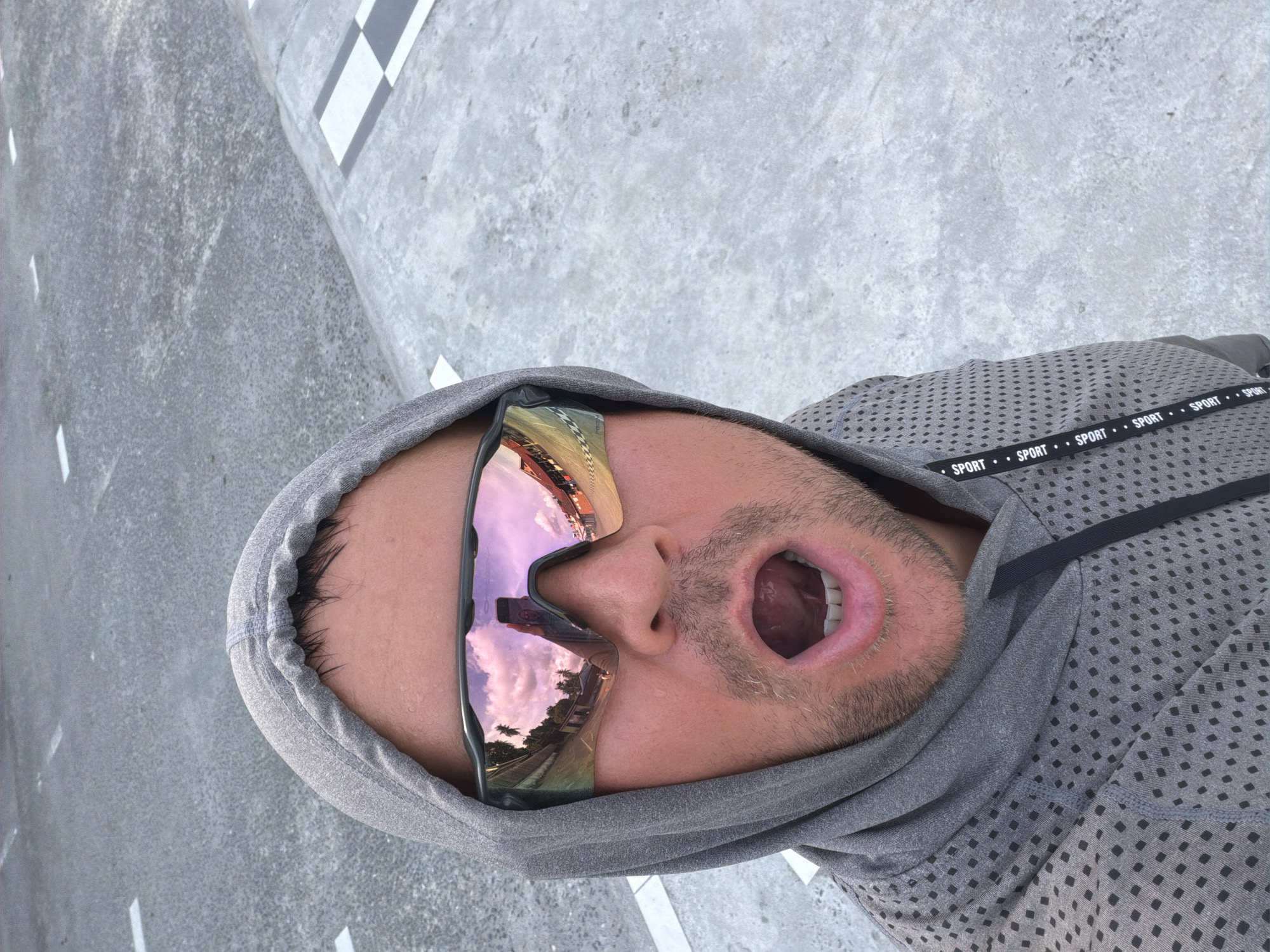 A man wearing a grey hooded jacket and performance sunglasses takes a selfie outdoors. The sunglasses reflect a sky with pink clouds and palm trees, and a building with red and yellow accents. The man's mouth is open in a surprised or shouting expression, and he has a light stubble on his face. The background appears to be a paved surface with white lines, suggesting a road or track. There is also a checkered pattern on the ground in the upper right corner. The location is Baan Mae Nam, Thailand.