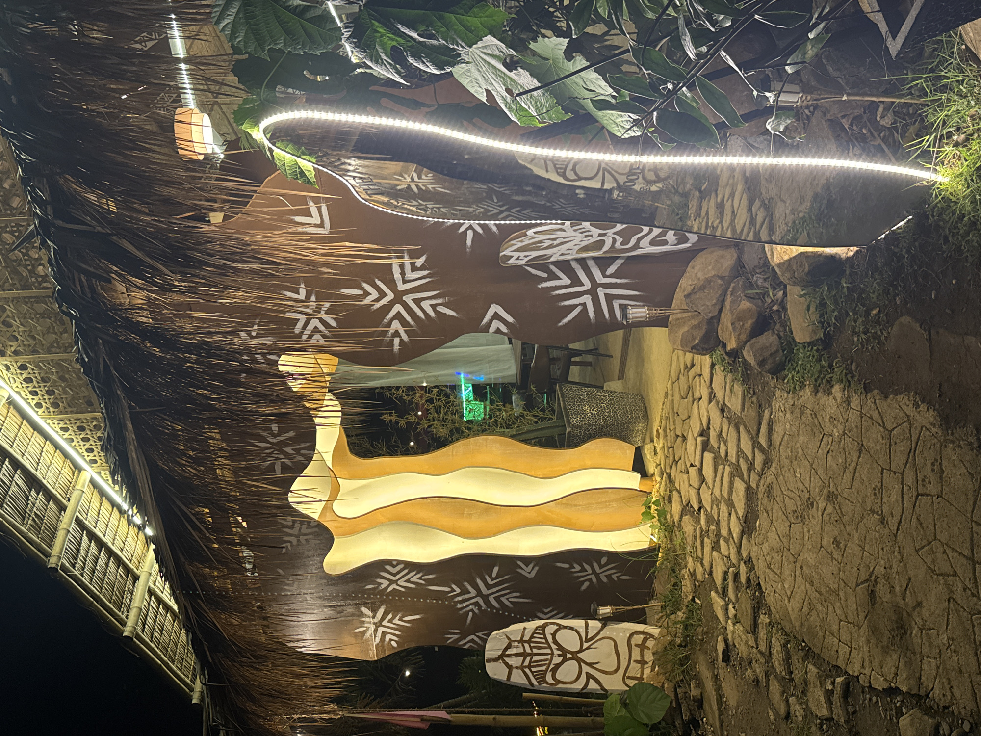 This photograph, taken at night in Davao City, Philippines, shows a structure with a thatched roof on the left, partially obscuring the sky. The roof is decorated with white geometric patterns and what appears to be a white painted tiki mask. A string of white LED lights runs along the edge of the roof. To the right of the thatched structure is a wall with wavy, tan-colored panels. Beyond the wall, glimpses of the interior reveal a chair with a leopard print cushion and green plants. The right side of the image shows a stone pathway or wall overgrown with grass and other vegetation. A bamboo structure is visible on the left.