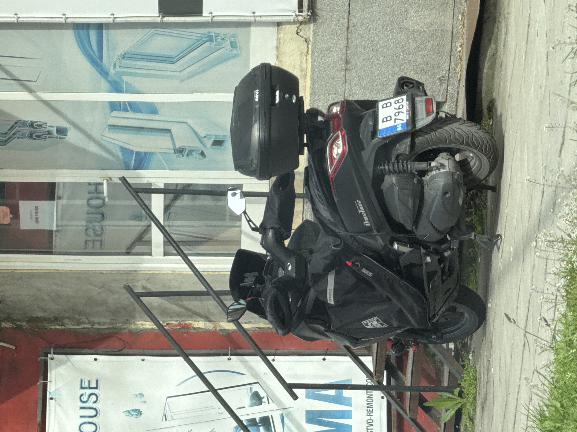 A black scooter with a GIVI top case is parked on a sidewalk in front of a building. The scooter has a Bulgarian license plate that reads "BB BG 7968." The scooter has bags on it and is partially obscured by metal scaffolding. The building has a storefront window displaying window or door frame components. A red painted wall and banner are visible below the window. The banner has the partial words "OUSE" and "MA" on it, as well as "STVO-REMONT CO." A phone number "0806 316 933" is seen in the window. Green grass is visible to the right of the sidewalk. The location is likely in Varna, Bulgaria, due to the Bulgarian license plate and the Cyrillic letters on the banner and license plate.