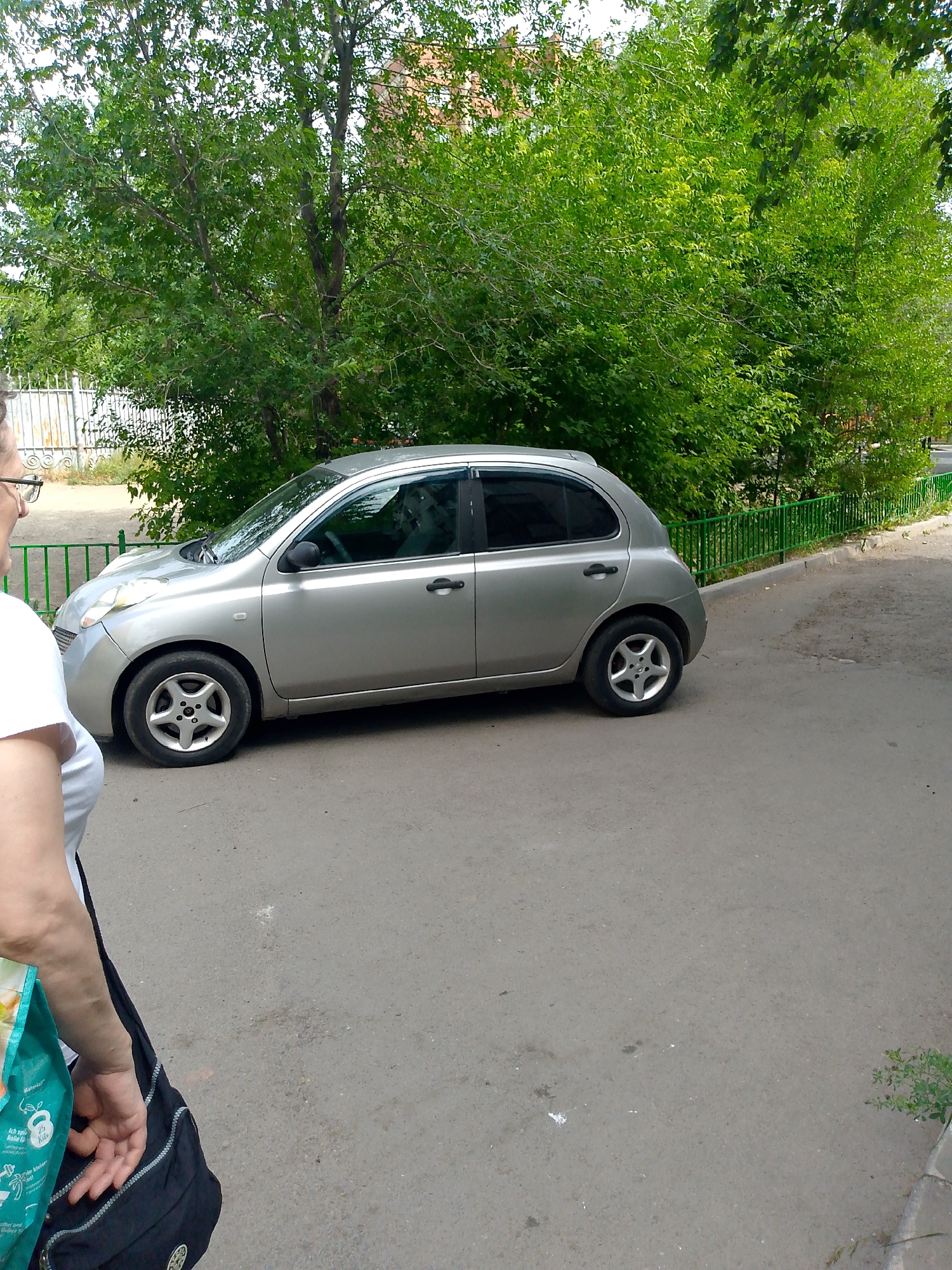 A medium-shot, eye-level, outdoor photograph taken on a sunny day shows a silver Nissan Micra parked on an asphalt surface, likely a residential courtyard or street. The car is facing left in the frame, showing its driver's side. A person, partially visible in the foreground from the chest down, is facing the car. The person is wearing a white t-shirt and has a black shoulder bag draped over their arm, with a green reusable shopping bag partially visible near their hand.
Behind the car, lush green trees with dense foliage dominate the background, suggesting a warm climate and summertime. A bright green metal fence runs horizontally behind the car and trees, separating the asphalt area from a patch of grass or unpaved ground. A hint of a building with reddish-brown brickwork is visible behind the trees on the left side of the image. The asphalt surface shows some minor wear and tear, with scattered debris.
The overall impression is a mundane, everyday scene, possibly in a city neighborhood. Based on the architectural style of the fence and the surrounding vegetation, and considering the style of the car which was commonly sold in the region, the location can be inferred as Astana, Kazakhstan, likely in a residential area with mature trees. The image captures a moment of pause, with the person seemingly observing or interacting with the parked vehicle.