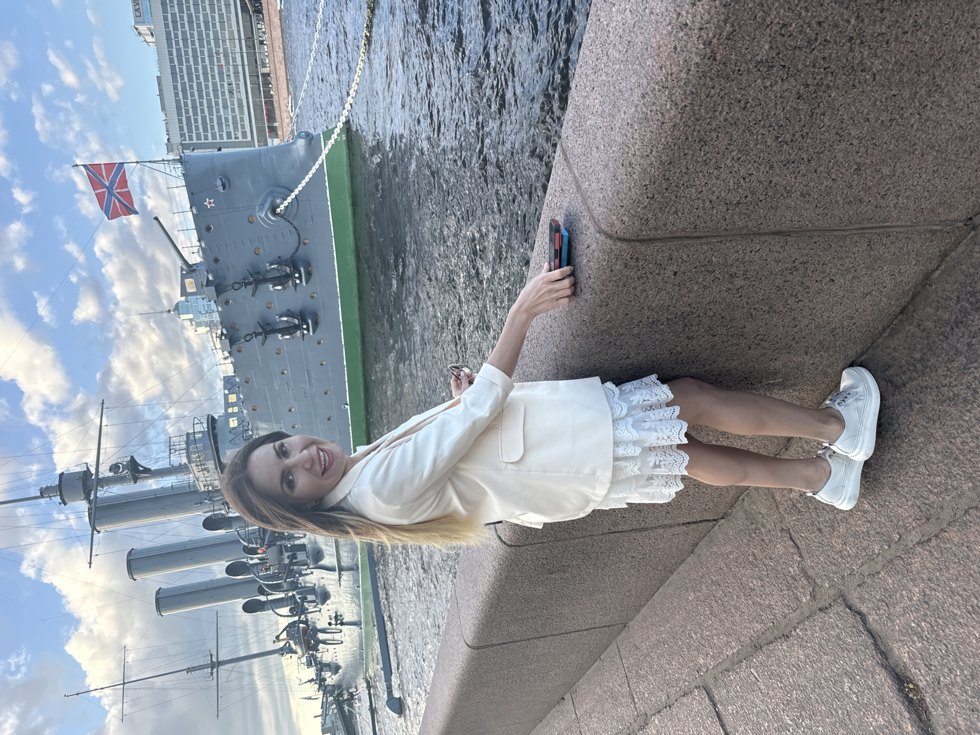 A woman stands on a stone embankment overlooking a river. Behind her, the Aurora cruiser, a museum ship and historic landmark, is docked. The ship is painted grey with a green hull and flies the Russian Naval Ensign. The woman is wearing a white blazer over a white lace dress and white sneakers. She is holding a mobile phone in her right hand and smiling at the camera. In the background, buildings of Saint Petersburg can be seen. The sky is partly cloudy with the sun setting.