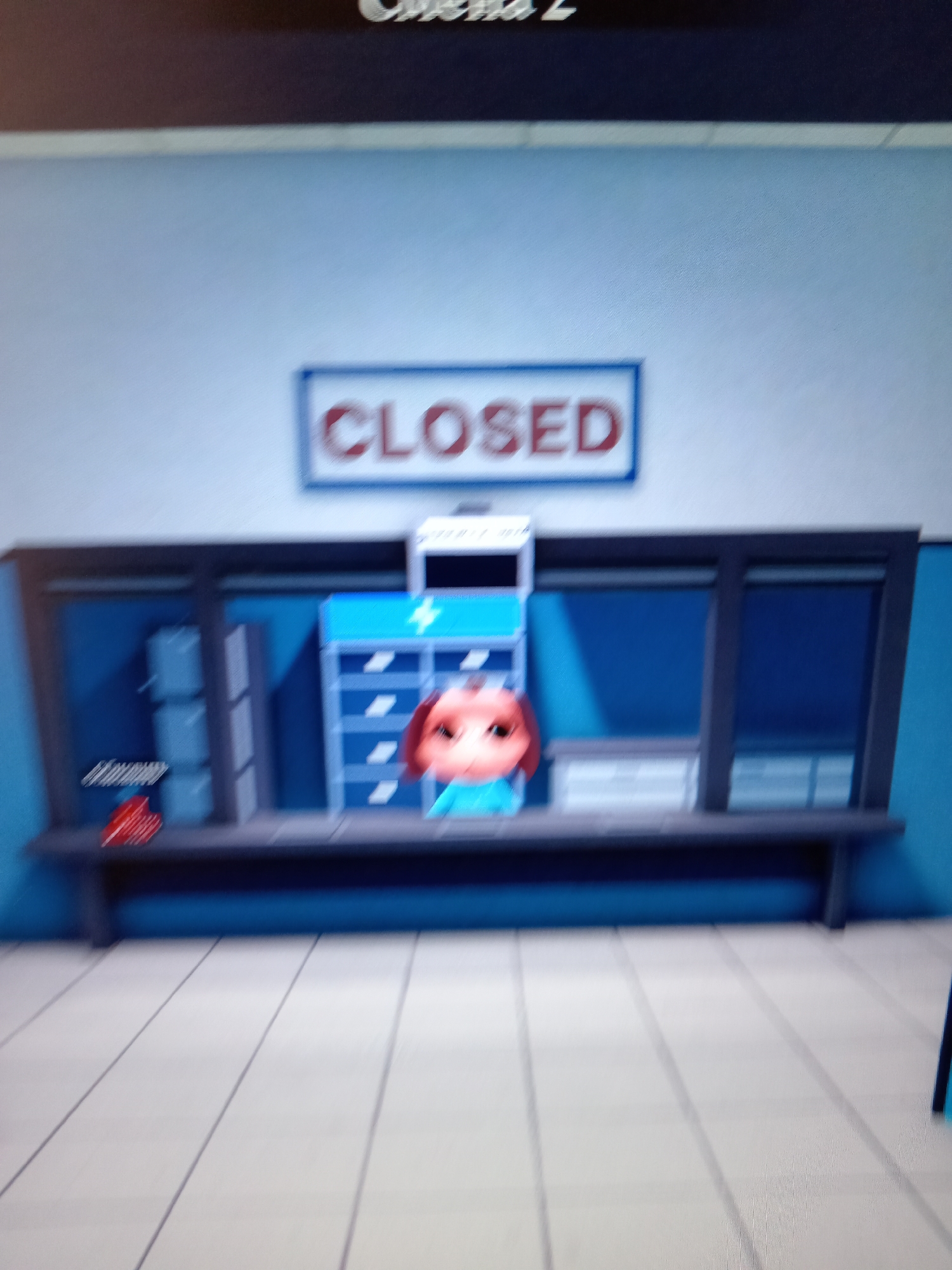 A video game screenshot shows a brightly lit, sterile environment with a counter and a "CLOSED" sign above it. A cartoonish character with pink skin and big eyes, wearing a blue shirt, is visible behind the counter. To the left of the character, there are several shelves stocked with what appear to be gray boxes, and a red object is on the counter in front of them. The floor is tiled in a light color. The image is blurry and appears to be taken from a screen, suggesting it is not a real-world scene. There is no information in the image that would indicate a specific location in the city of Rudny, Kazakhstan.