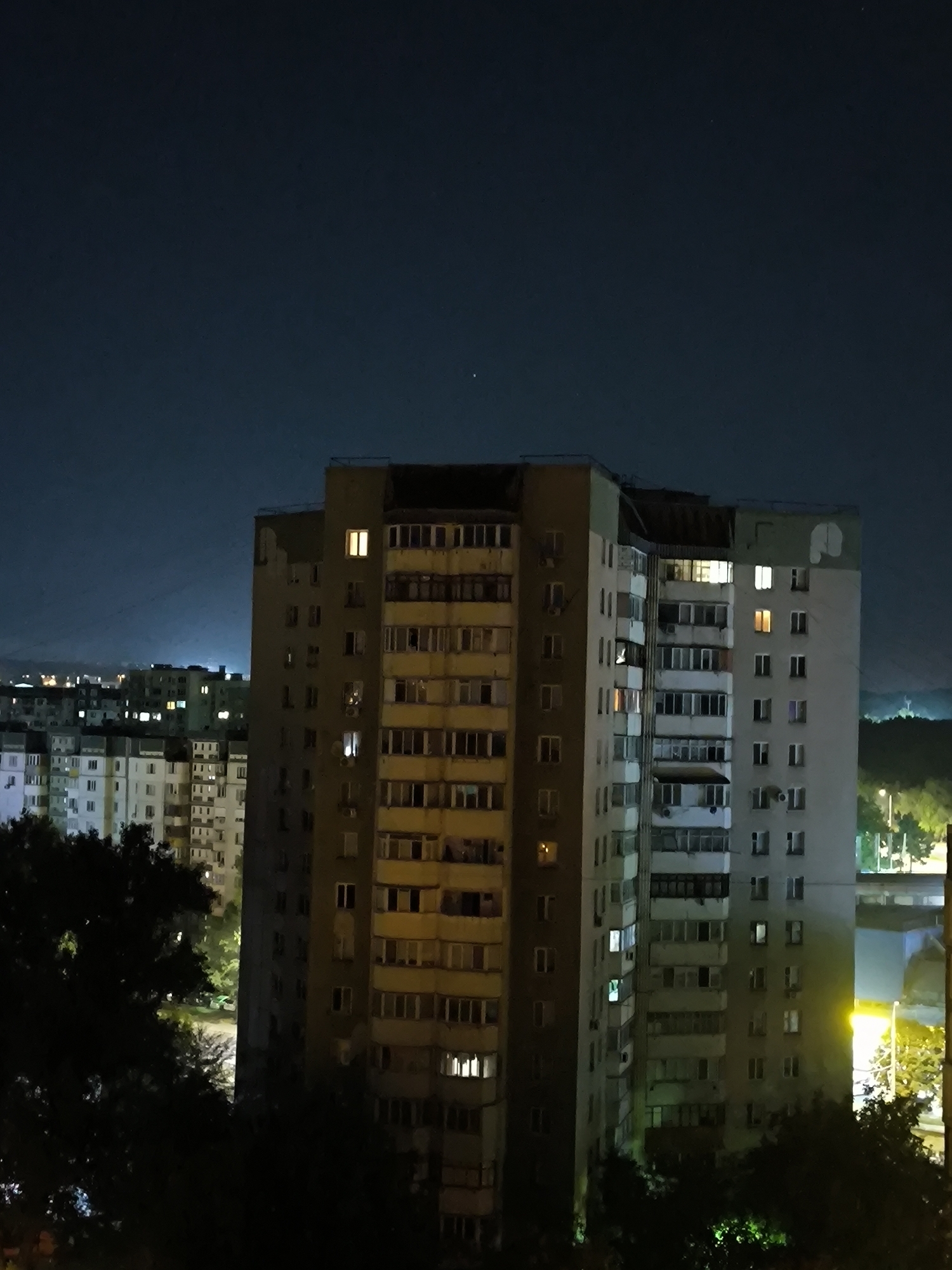 The image shows a night view of residential buildings in Chișinău, Moldova. The foreground is dominated by the dark silhouettes of trees, partially obscuring the view of the buildings. Behind the trees, several multi-story apartment buildings are visible. The building in the center of the frame is tall and appears to be made of concrete, with numerous windows. Some of these windows are illuminated, indicating occupancy. To the left and behind this central building are other apartment blocks of varying heights and colors, with more lit windows scattered throughout. In the background, a faint outline of distant hills or mountains can be seen against the dark night sky, which has a slight blueish tint. A few lights from streetlamps or other structures are visible in the far distance, and a bright yellow light is emanating from the right side of the frame, likely from a streetlamp or building exterior. The overall impression is a typical urban night scene in a city.
