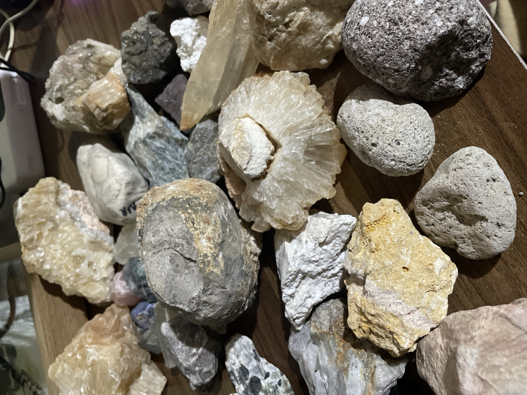 The image provides an overhead, close-up view of a diverse collection of rocks and minerals displayed on a warm-toned wooden surface. The collection features a variety of geological specimens differing in size, shape, color, and texture.
Prominent among the minerals are several crystalline formations. A large, elongated, translucent pale yellow crystal, possibly quartz or calcite, is visible towards the upper center-left. Below it, a distinctive circular, fan-like cluster of white to pale yellow crystals, reminiscent of aragonite or fibrous calcite, forms a focal point. Other crystalline rocks include rough, translucent pieces in shades of pale yellow and white, some showing distinct facets.
Interspersed with the crystalline specimens are various types of rocks. These include dark grey, speckled igneous rocks, such as a large rounded one in the upper right. Several smooth, light grey, and somewhat porous rocks, likely weathered river stones or volcanic rocks, are present in the mid-right. There are also rough, earthy-toned rocks in browns, grays, and yellowish-beige hues, some with visible textures and possibly mineral inclusions. Smaller, multicolored pebbles in pink and blue are subtly visible nestled among larger rocks in one area.
On the far left, a white electronic device, possibly a power bank or charger, with a black cable, is partially visible, indicating an indoor setting. The wooden surface appears to be a table or shelf, suggesting the rocks are part of a personal collection or display rather than in their natural environment. The lighting is artificial, casting subtle shadows that highlight the textures and contours of the rocks. Faint, indecipherable block letters, possibly "MM R" or similar, are visible on a light grey rock towards the upper left quadrant of the full image. No specific activity is depicted; the image is a still life of the rock collection.