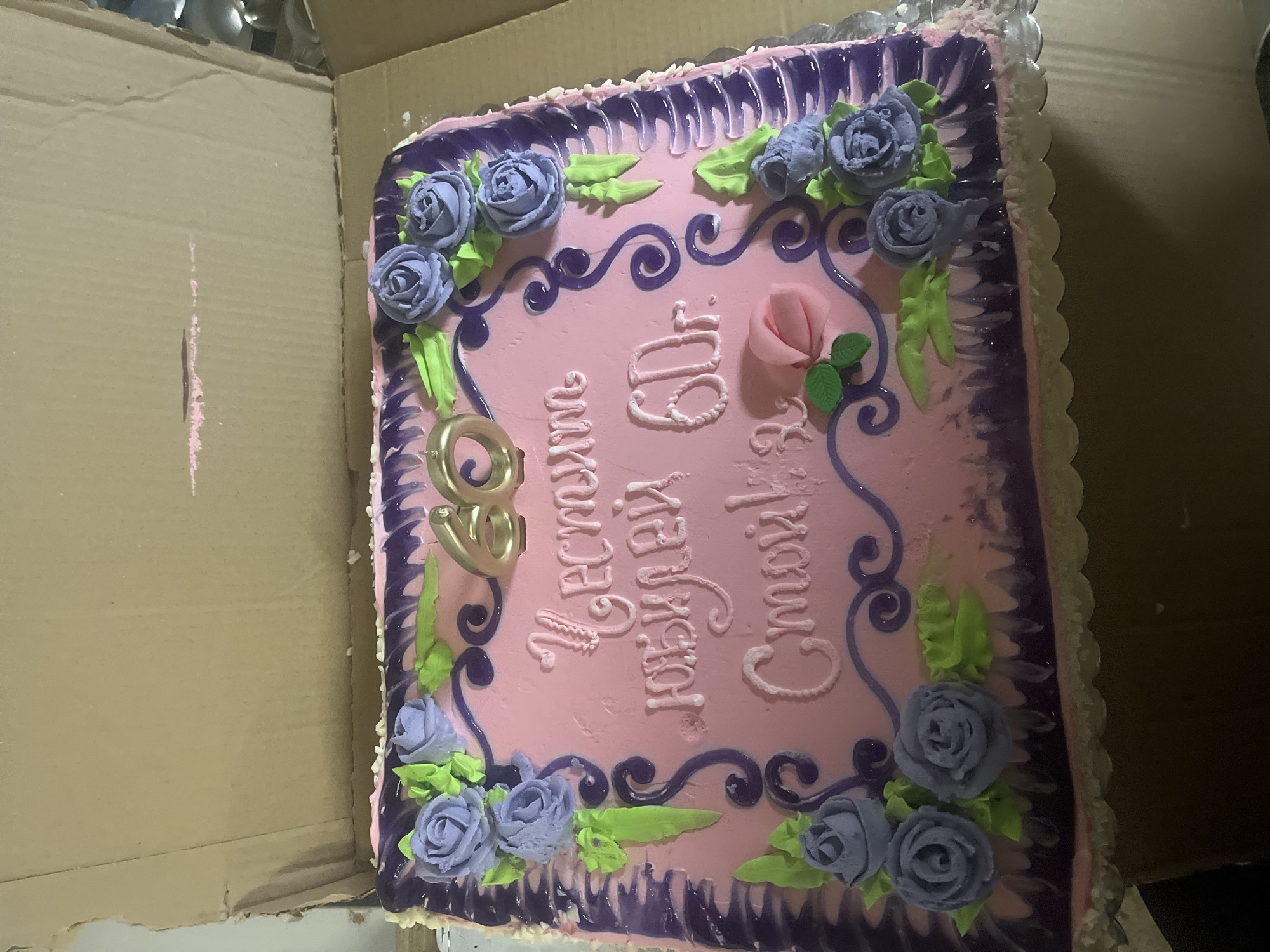 The image shows a square birthday cake in a cardboard box. The cake has pink frosting with purple decorative borders and stylized purple roses with green leaves along the edges. "60" is rendered in gold-colored candles. White lettering in Cyrillic is inscribed on the cake. A single pink rose is located on the right side of the cake. The box appears to be made of corrugated cardboard. The location of Kosten, Bulgaria is not apparent from this image.