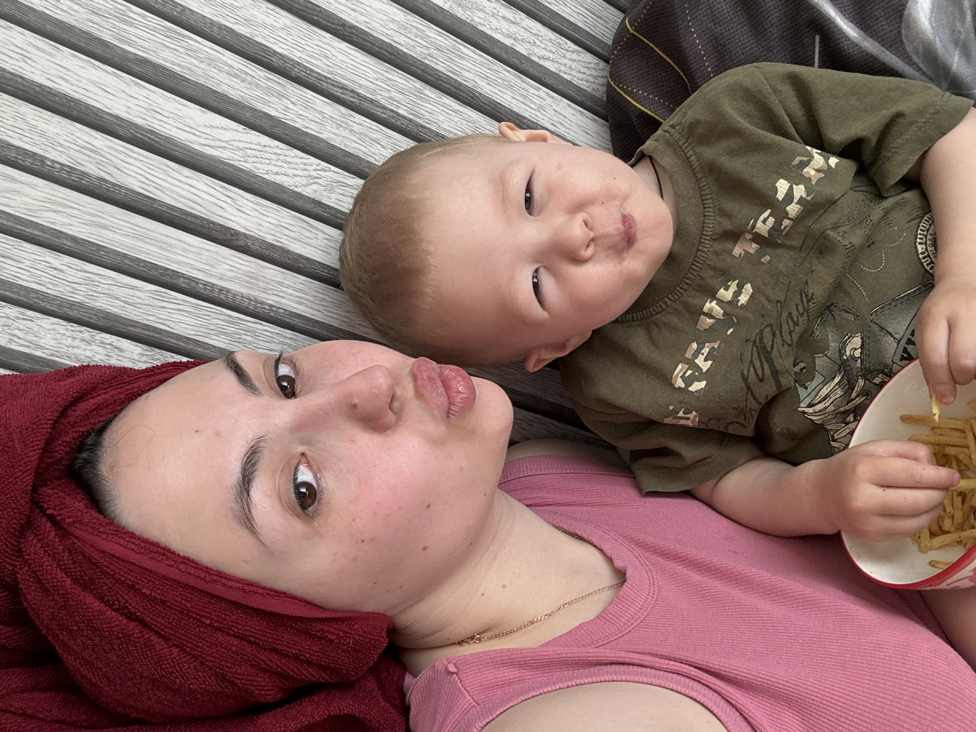 A woman with a red towel wrapped around her head and wearing a pink shirt lies on a horizontally striped surface next to a toddler who is wearing a green shirt and holding a bowl of french fries. This photo was taken in Марковский, Russia.