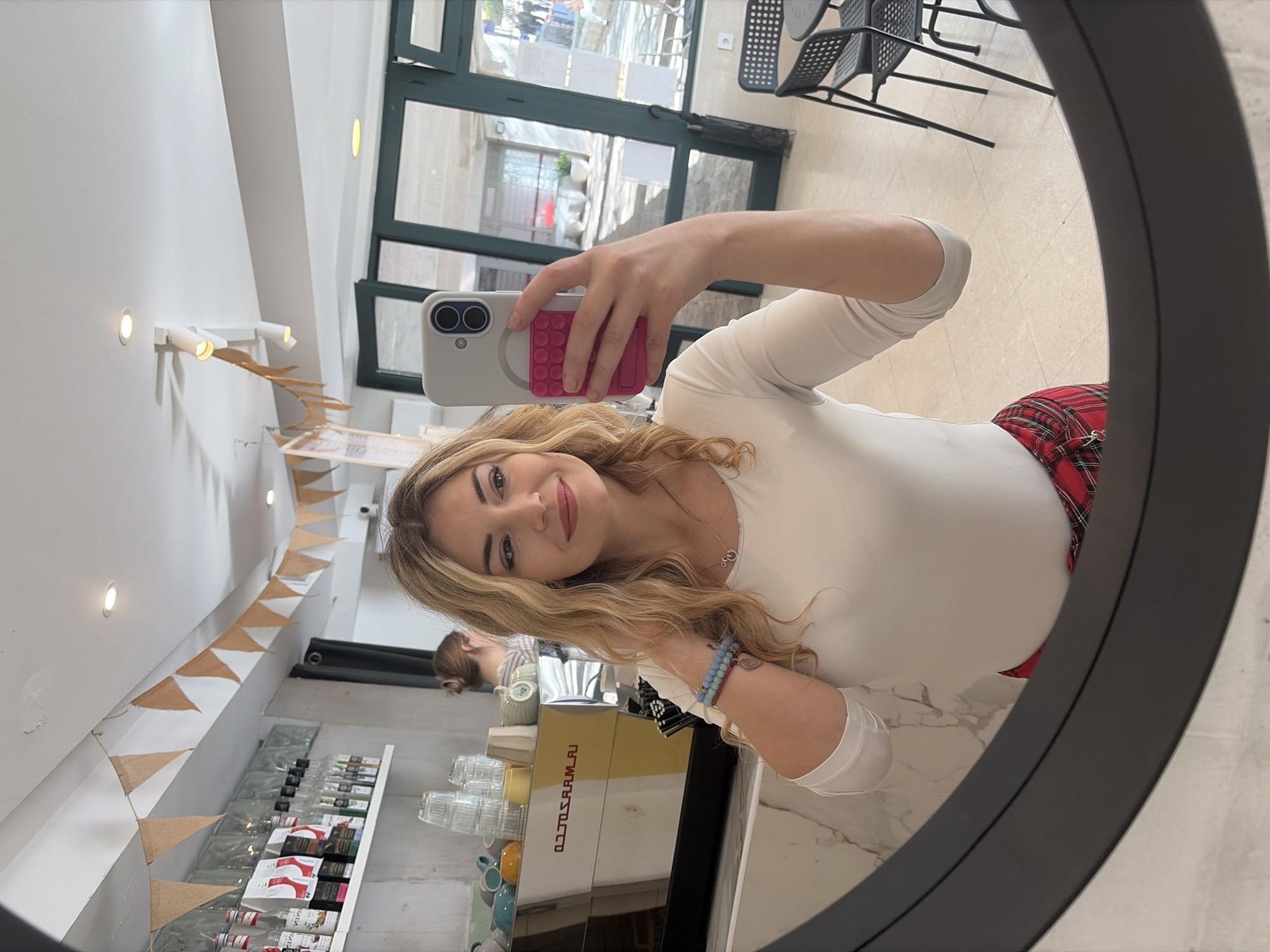 A young woman takes a selfie in a round mirror. She is wearing a white long-sleeve top and a red plaid skirt. She is holding a phone with a light grey case and a pink grip. In the background, a coffee shop bar area is visible with bottles, coffee cups, and a coffee machine. A woman with a bun is visible behind the bar. A doorway leads to an outdoor seating area with black metal chairs. A string of pennant banners is strung across the ceiling. The location is likely a cafe in Budva, Montenegro.