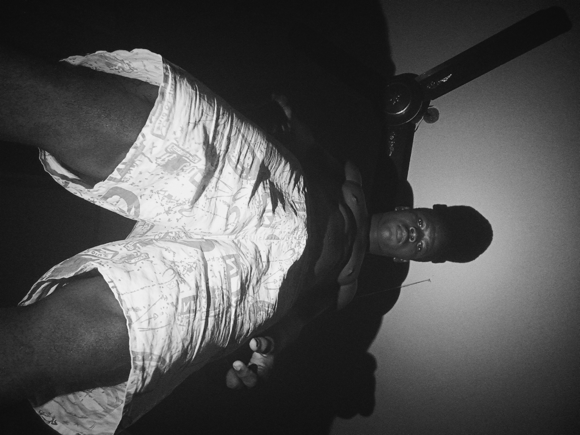 A low-angle, black and white photograph depicts a person wearing patterned shorts lying down, positioned at an angle relative to the camera, with a ceiling fan visible in the upper right corner. The person is a dark-skinned male with a close-cropped haircut. The photograph is taken indoors, with a wall as the background. The location, Nkpor, Nigeria, is not directly evident from the image itself.