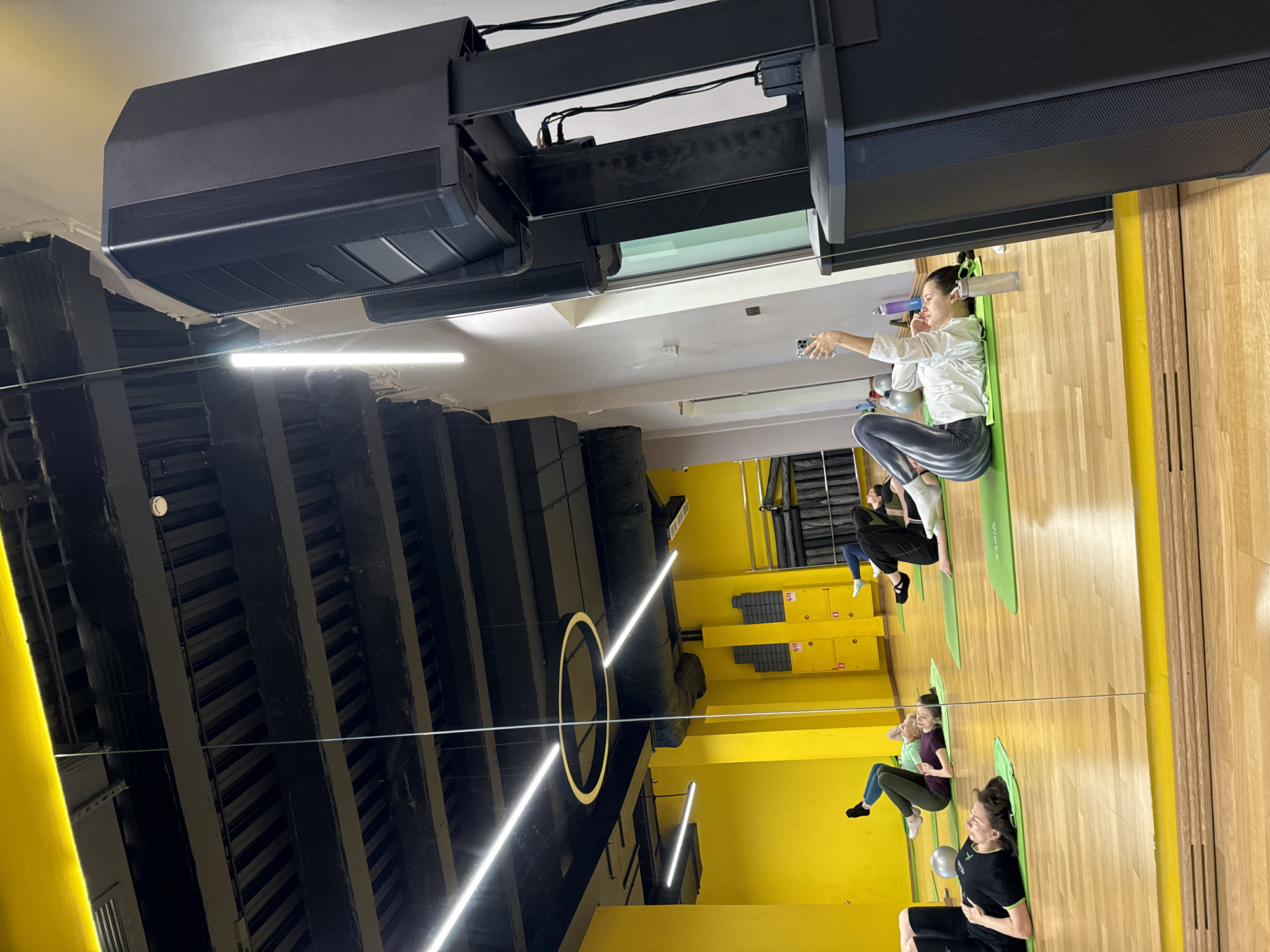 An indoor scene captures a group of women participating in a fitness class, likely yoga or Pilates, within a studio at Vniissok, Russia. The image, taken from a low angle, features a prominent mirrored wall reflecting the room and its occupants, creating a dynamic visual effect.
In the foreground, a woman wearing a white long-sleeved shirt and grey patterned leggings sits on a green yoga mat, holding a smartphone in her outstretched arm, possibly taking a selfie or recording. Behind her, another woman in a black top and black leggings is seated on a similar green mat, with a small grey exercise ball nearby. Further into the room, reflections show at least two more women on green mats, all appearing to be engaged in the exercise session. One of the mats clearly displays the brand name "VIXIA".
The studio has a modern aesthetic with light wooden floors and bright yellow walls or pillars, contrasting with a dark, industrial-style ceiling featuring exposed black beams and large black speaker systems. Long, linear fluorescent lights illuminate the space, alongside a distinctive circular light fixture. The reflections in the mirror create multiple instances of these architectural features. The overall atmosphere suggests an active and focused environment for physical well-being.