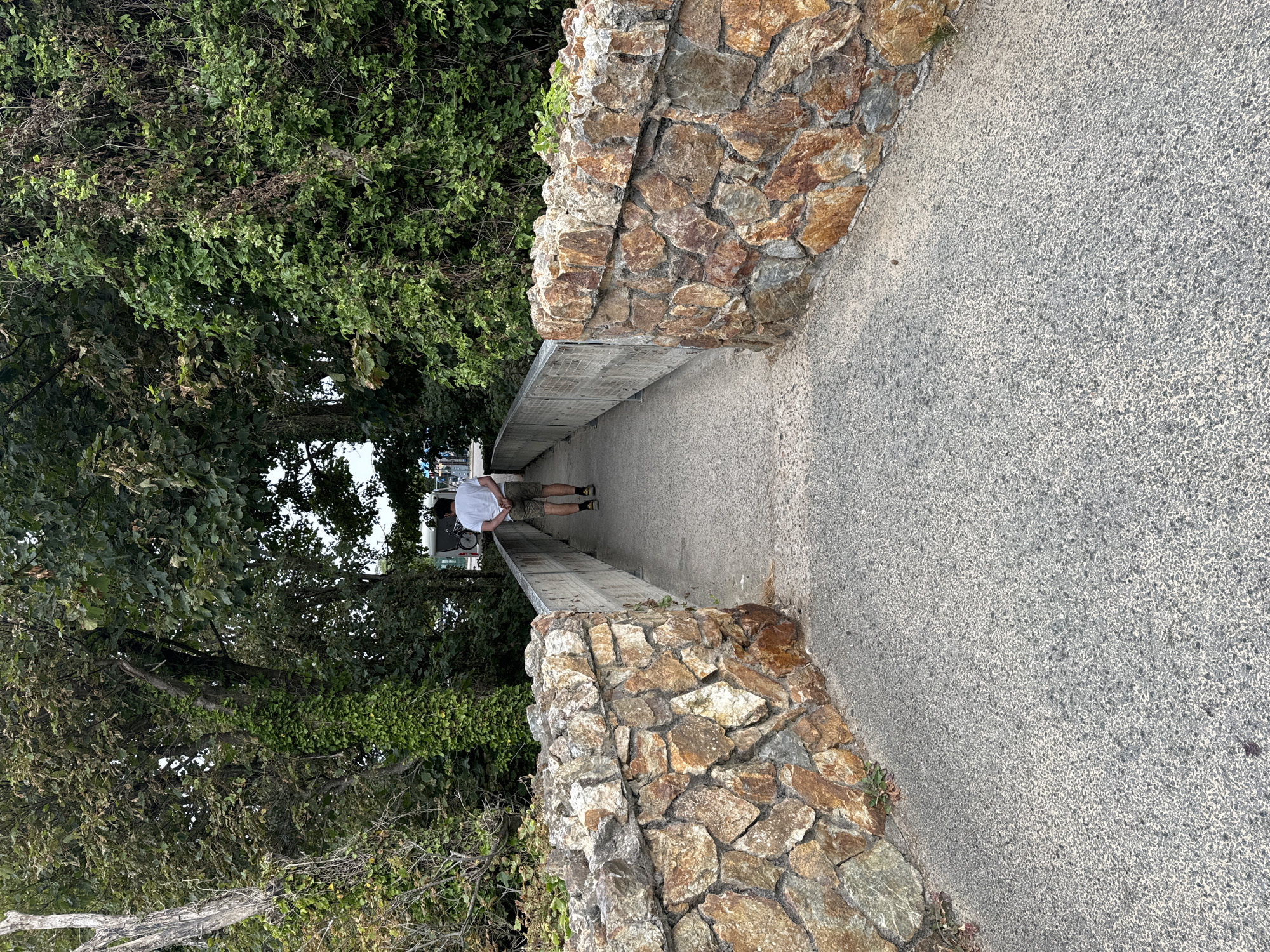 A person is walking away from the camera on a paved pathway that is flanked by stone walls on either side. The pathway leads towards a bridge with a metal railing. The stone walls are constructed from irregularly shaped, reddish-brown stones. The pathway is surrounded by dense green foliage and trees, suggesting a natural or park-like setting. The person is wearing a white t-shirt and khaki shorts, with their hands clasped behind their back. In the distance, a white van and a bicycle can be seen. The scene appears to be in Courtown, Ireland, possibly near a coastal area given the natural elements.